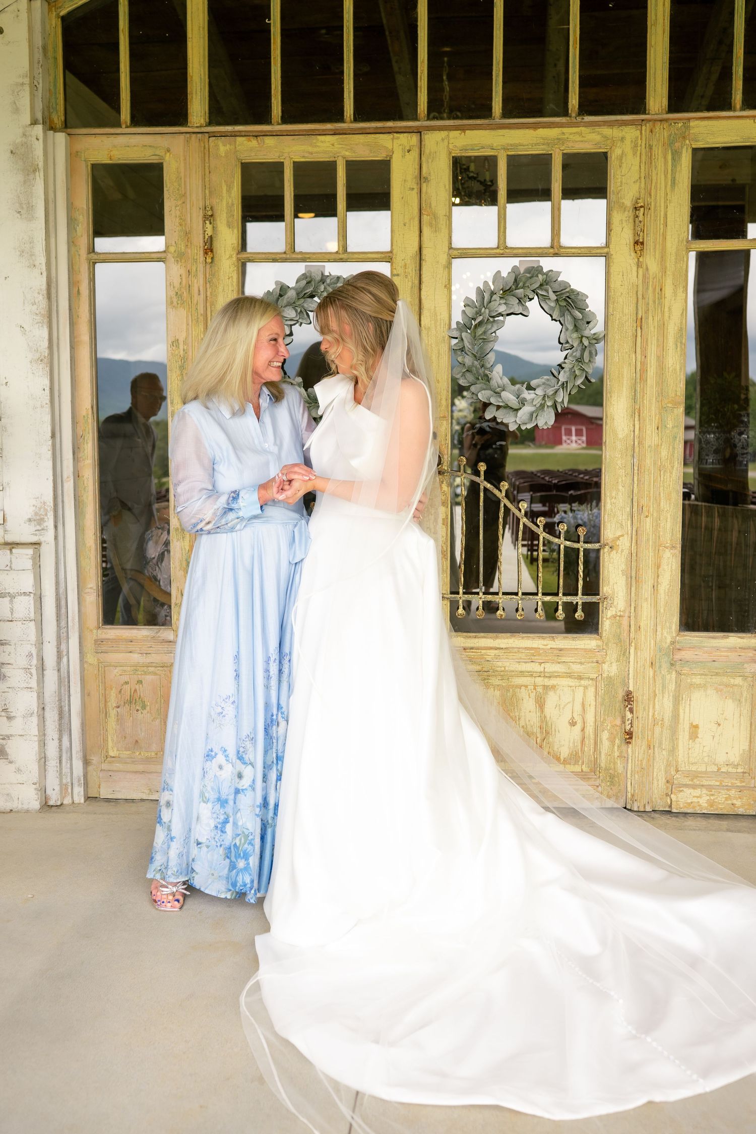 Bride in white gown with long veil shares tender moment with mother in blue dress.