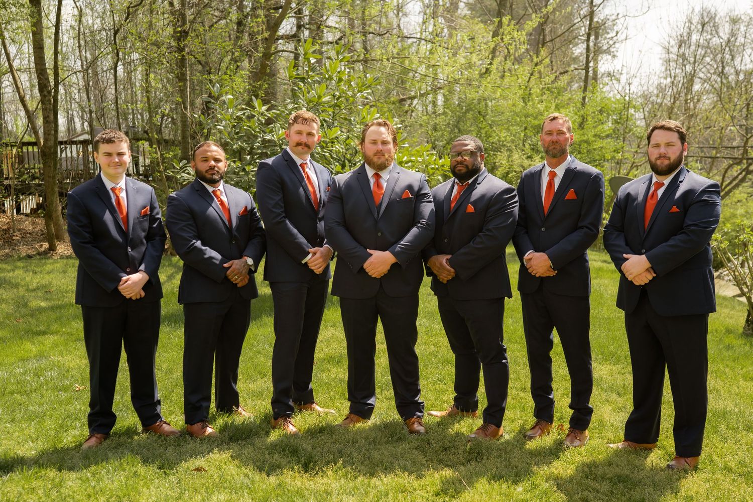 Seven groomsmen in navy suits with orange ties pose outdoors on green grass with trees in background.