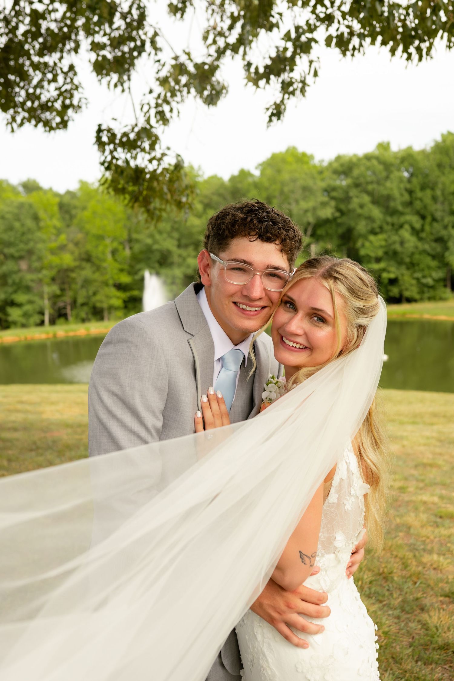 Bride and groom embracing outdoors near a pond, her veil flowing, wearing gray suit and white lace dress.