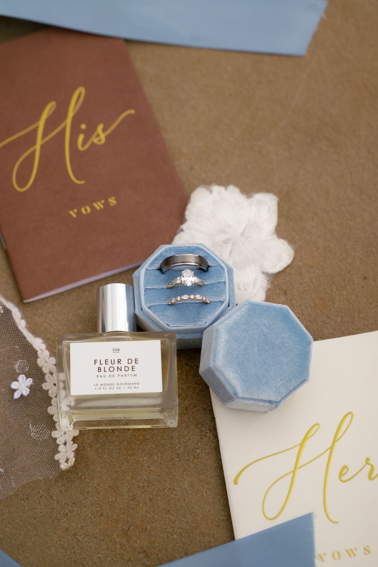 Blue velvet ring box with wedding bands, vow books, and Fleur de Blonde perfume in an elegant flat lay.