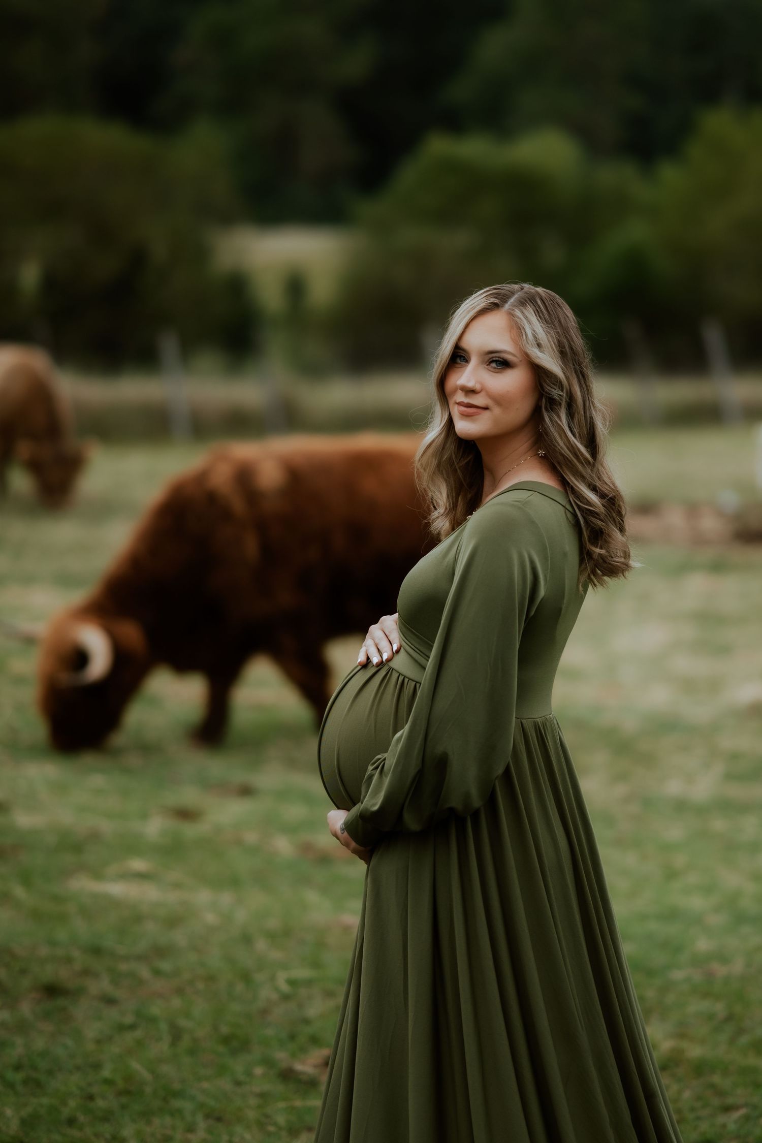 Pregnant woman in flowing olive dress poses in field with highland cows grazing.