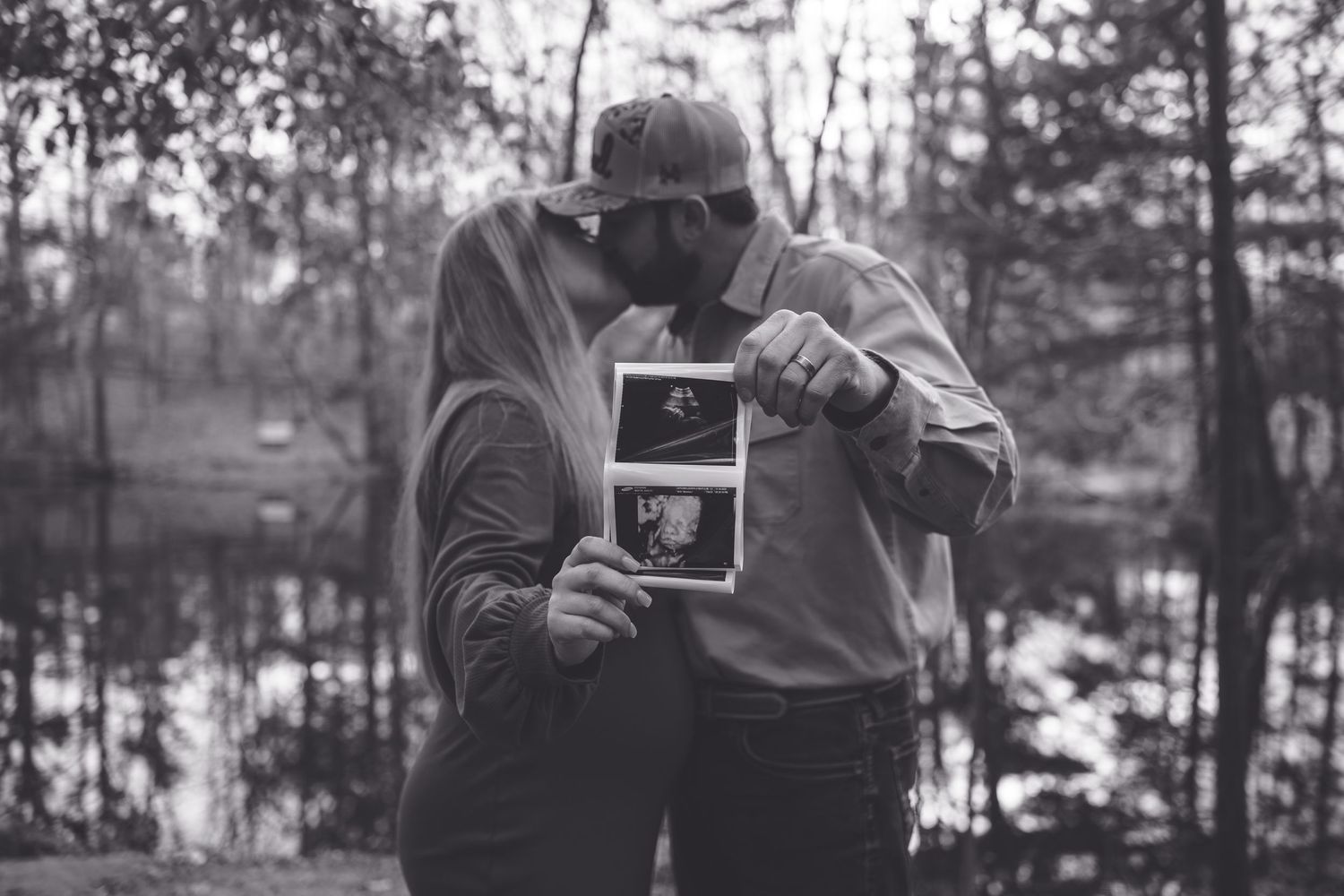 A couple shares a tender moment outdoors while holding an ultrasound photo in a black and white photograph.