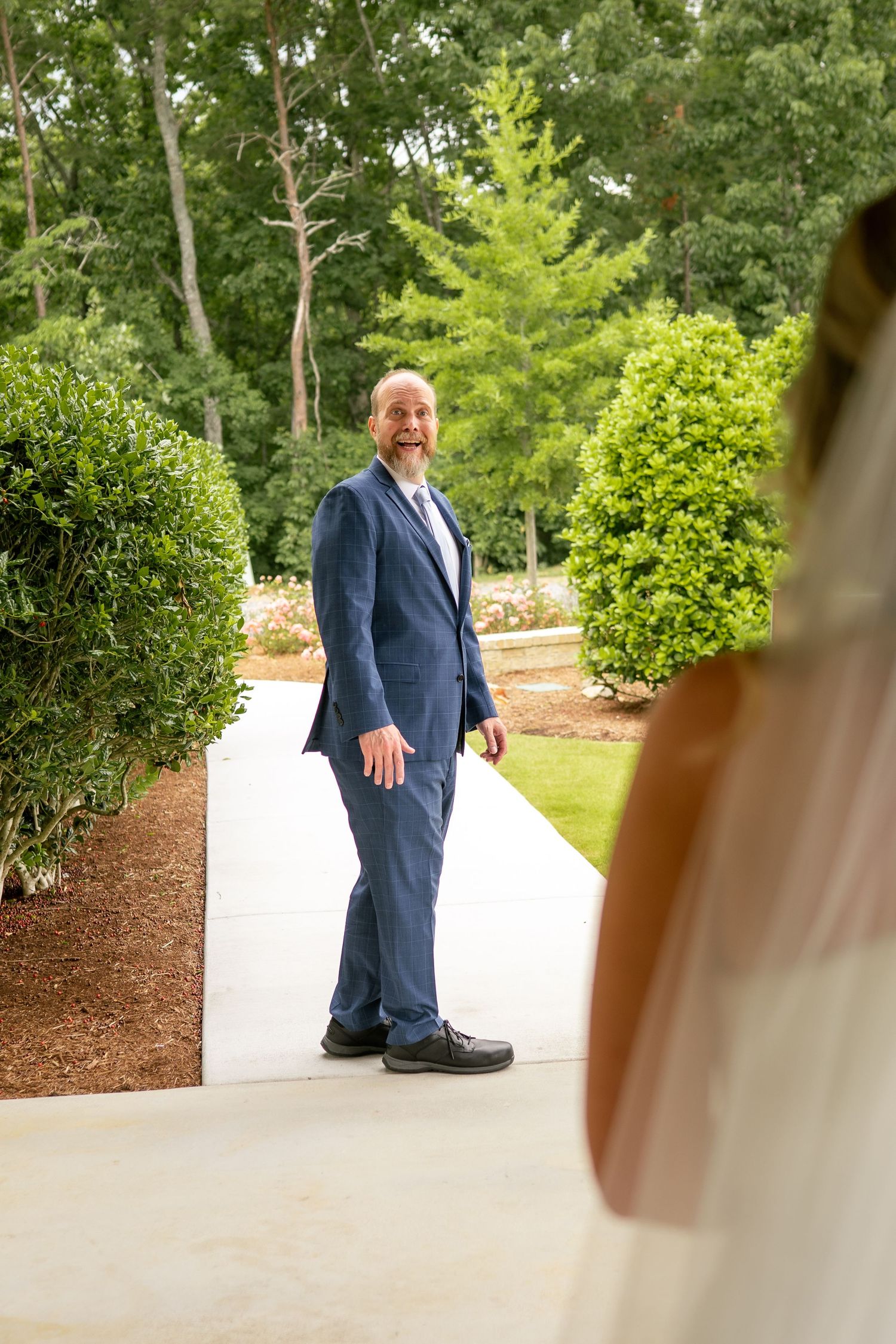 Smiling bald groom in navy suit turns to see bride during outdoor first look.
