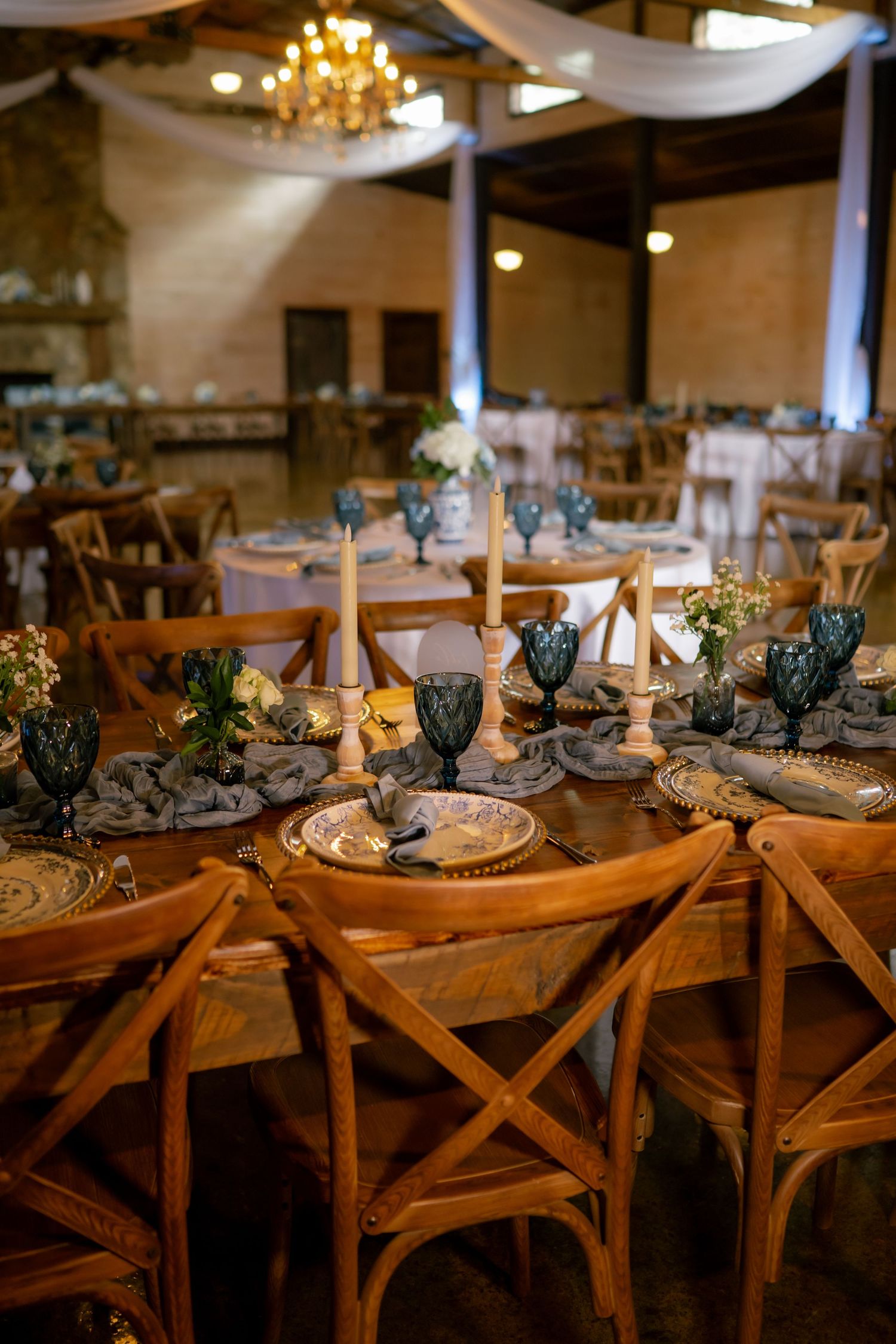 Elegant rustic wedding reception with wooden cross-back chairs, blue glassware, candlesticks, and floral centerpieces in a barn venue.