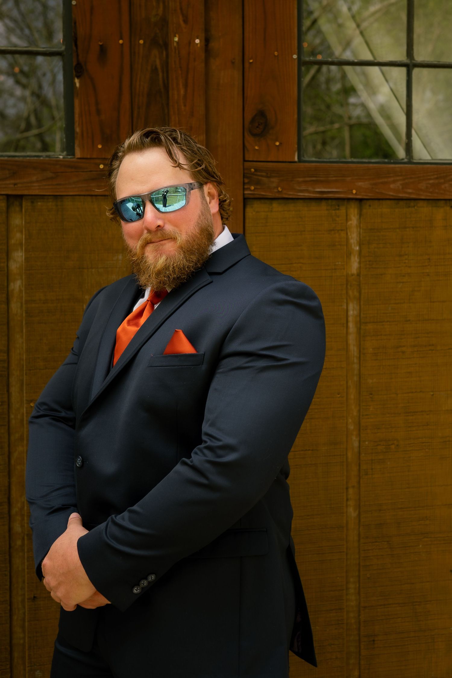 Bearded groom in navy suit with orange tie and pocket square wearing reflective sunglasses at outdoor wedding venue.