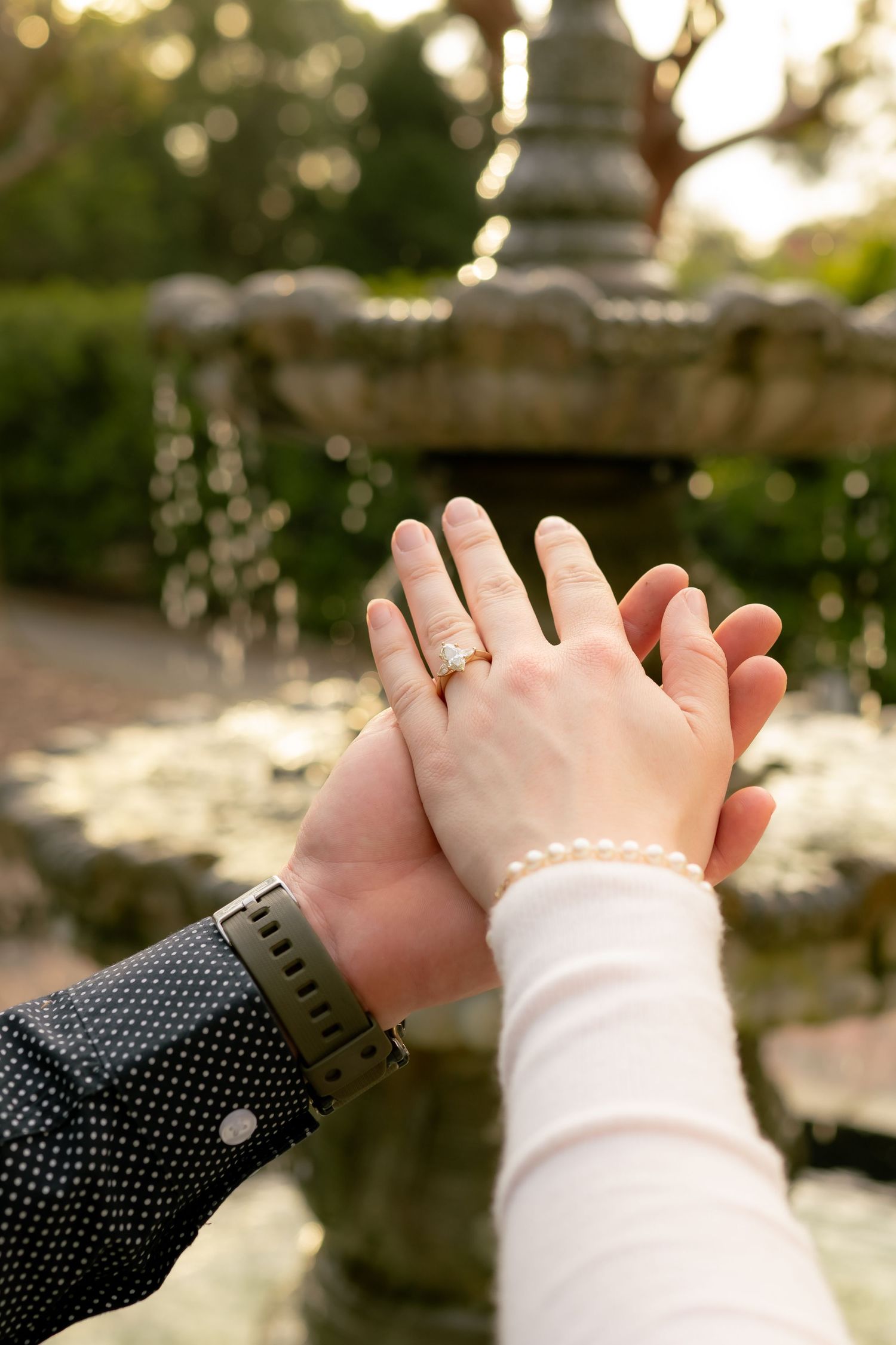Couple holding hands near a stone fountain, woman wearing an engagement ring and pearl bracelet, man in polka dot shirt.