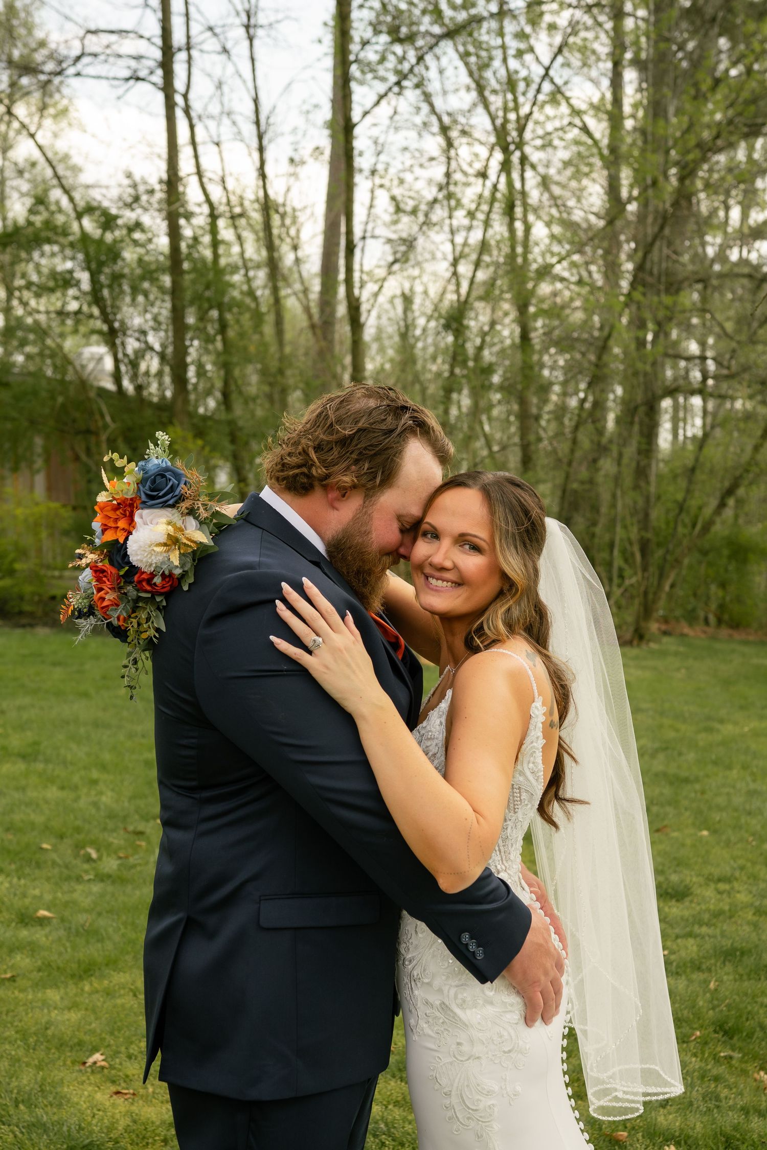 Smiling bride in lace gown with veil holds colorful bouquet while groom in navy suit embraces her outdoors.