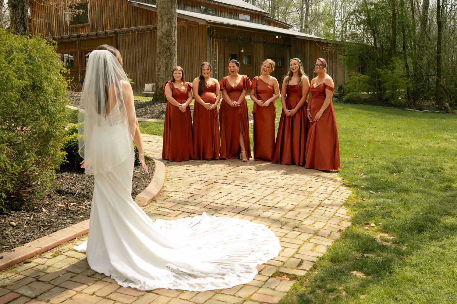 Bride in a lace veil and train reveals herself to bridesmaids in rust-colored gowns during an outdoor first look.