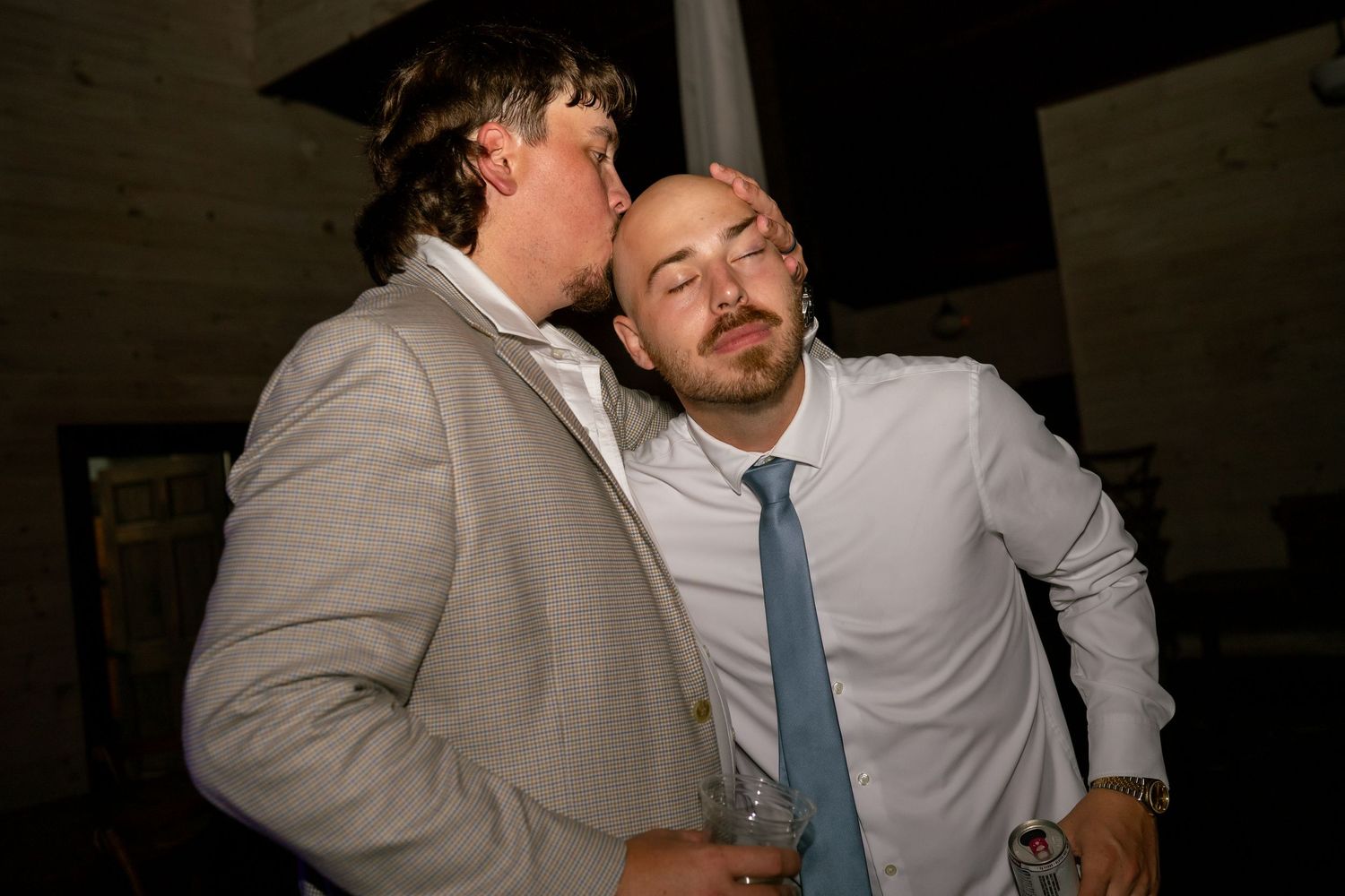 Two men in suits sharing a playful moment at a party, one kissing the other on the cheek while holding a drink.