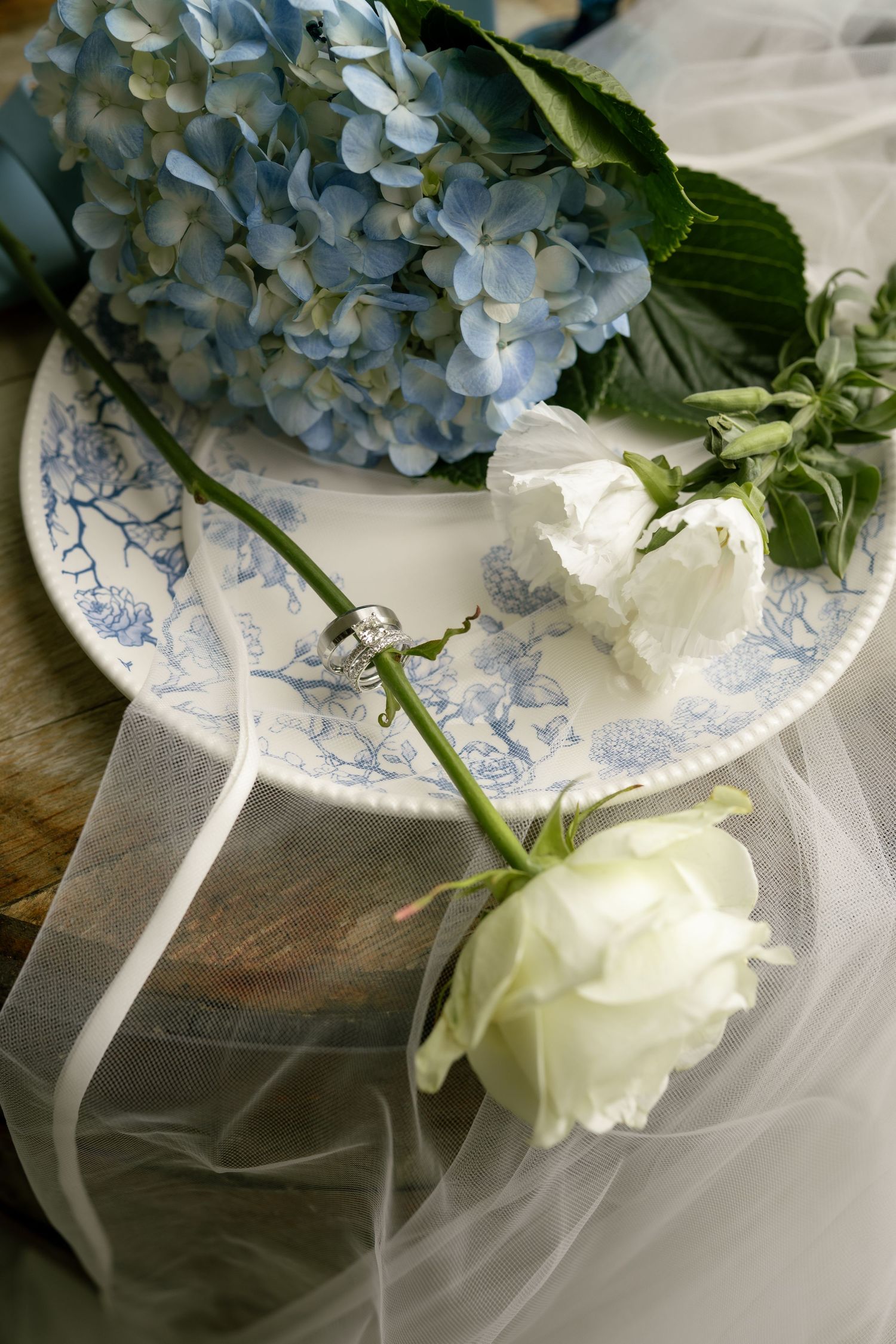 Blue hydrangeas and white roses with a diamond ring on a blue toile plate with sheer ribbon.