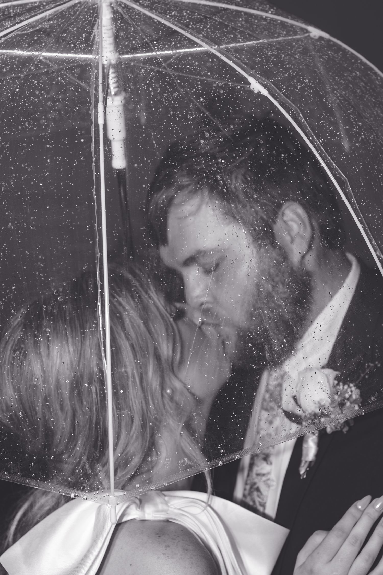 Bride and groom share a romantic kiss under a clear umbrella in the rain, black and white photo.