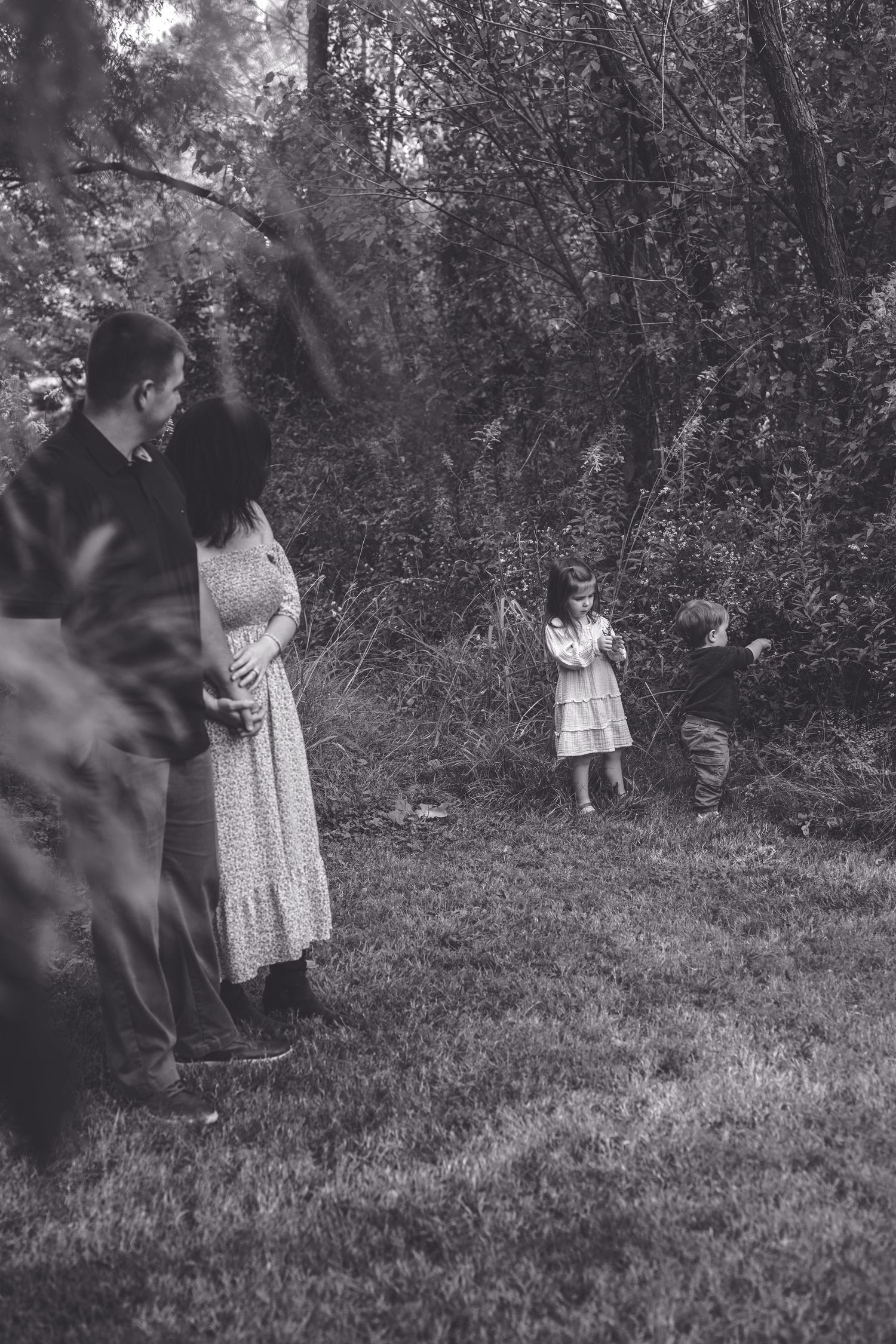 A vintage black and white photograph shows a family gathering in a wooded area with children playing in the background.