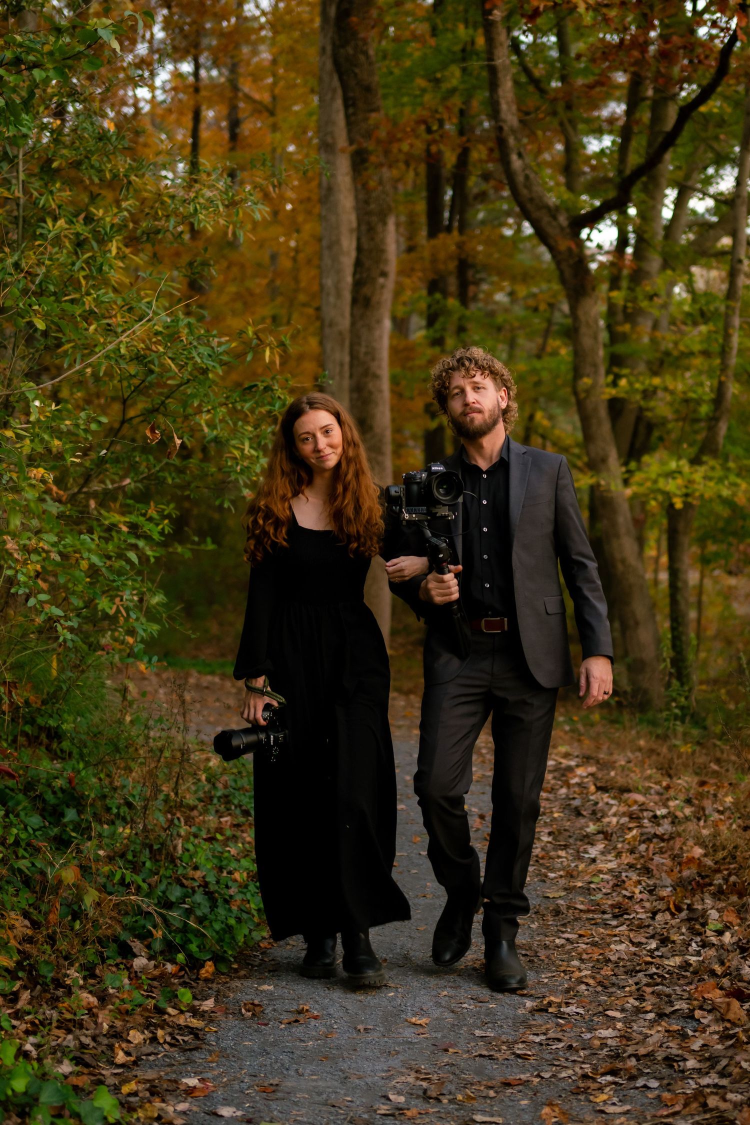 A Photographer and videographer walks together on a leaf-covered autumn forest path in moody lighting. Southern pines, NC