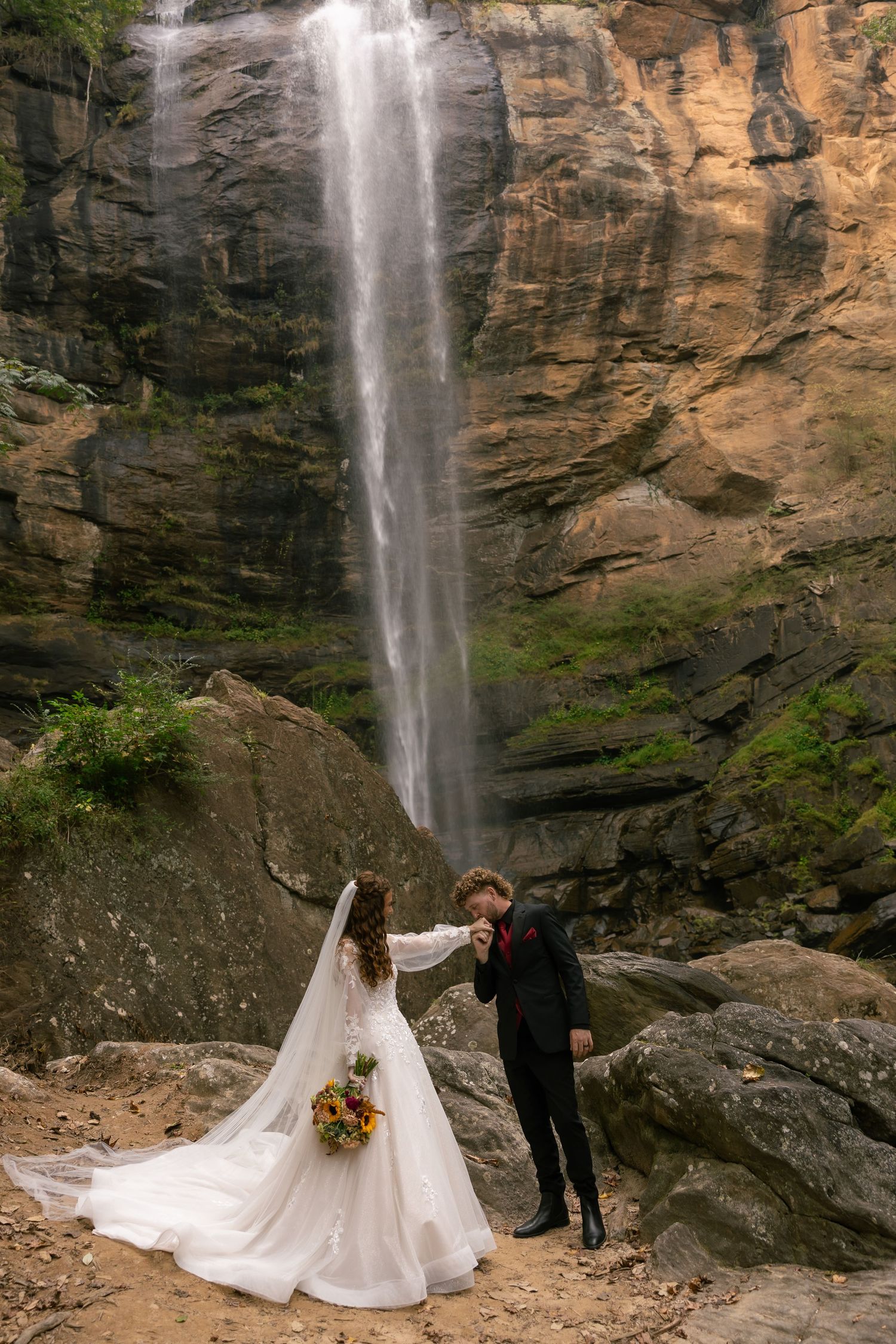 A couple in wedding attire poses in front of a majestic waterfall cascading down tall rocky cliffs. Toccoa falls, GA