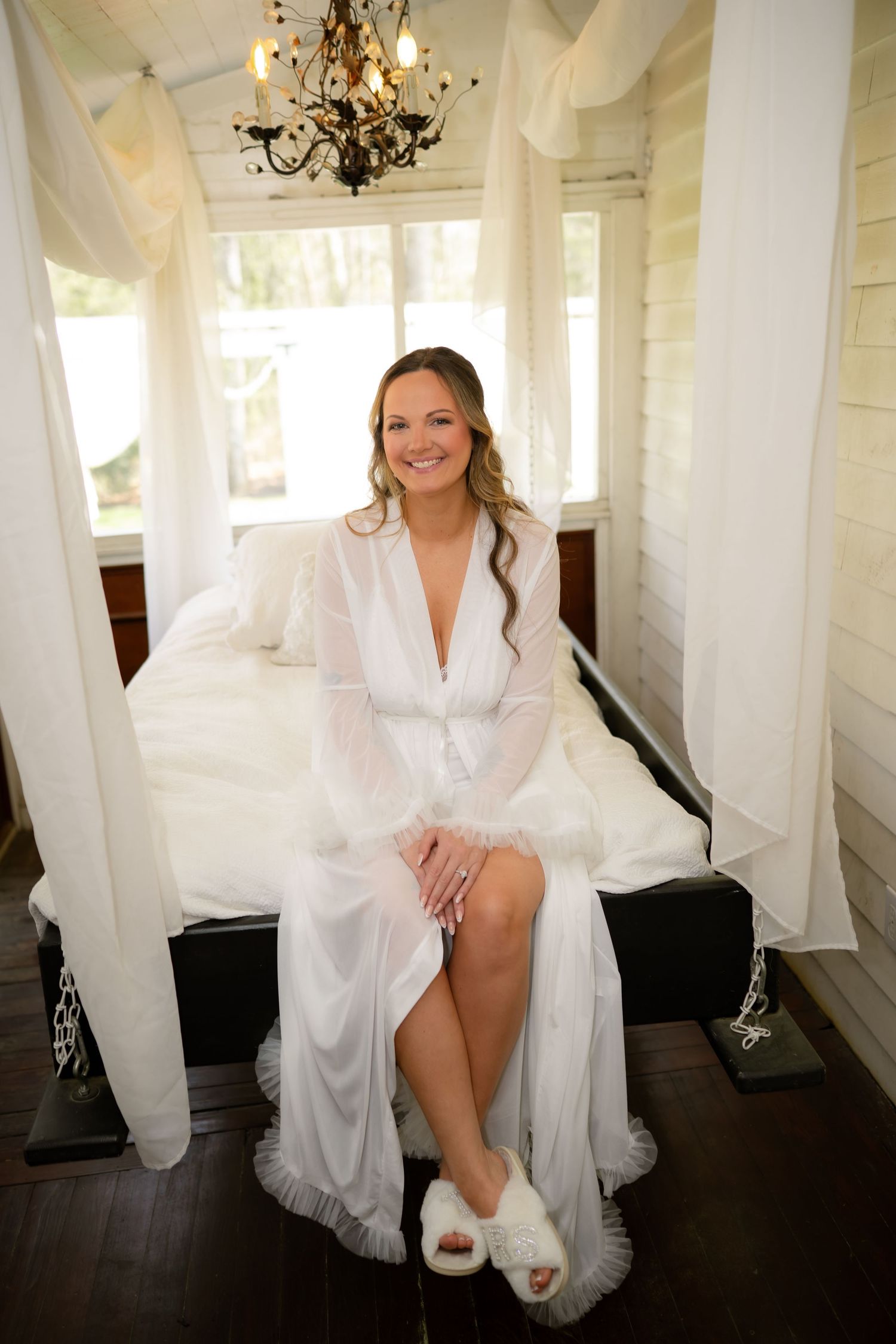 Smiling bride in white sheer robe and fluffy slippers sits on a canopy bed under a chandelier.
