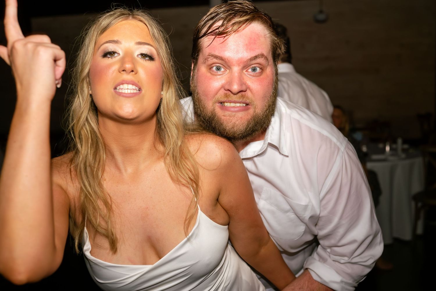 A woman in a white dress and a bearded man in a white shirt pose playfully at a party event.