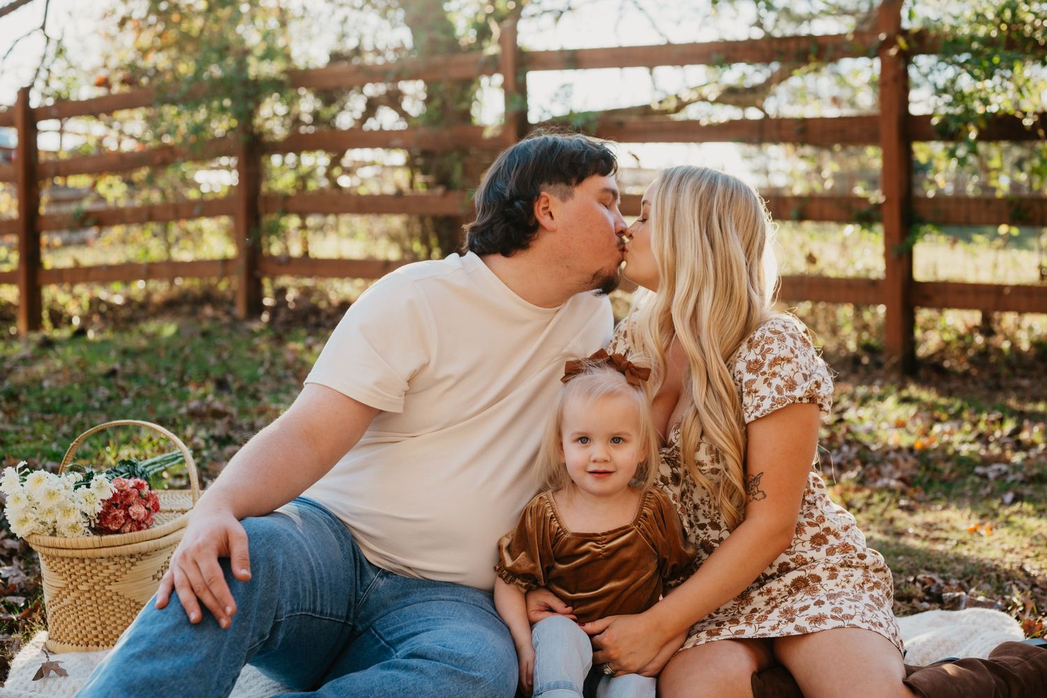 A couple shares a tender picnic moment with their toddler in a rustic outdoor setting during golden hour.
