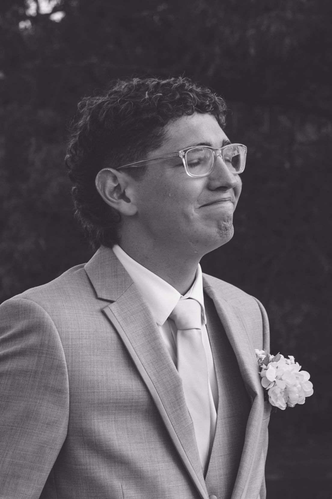 Young groomsman in gray suit with boutonniere and glasses smiling at wedding ceremony.