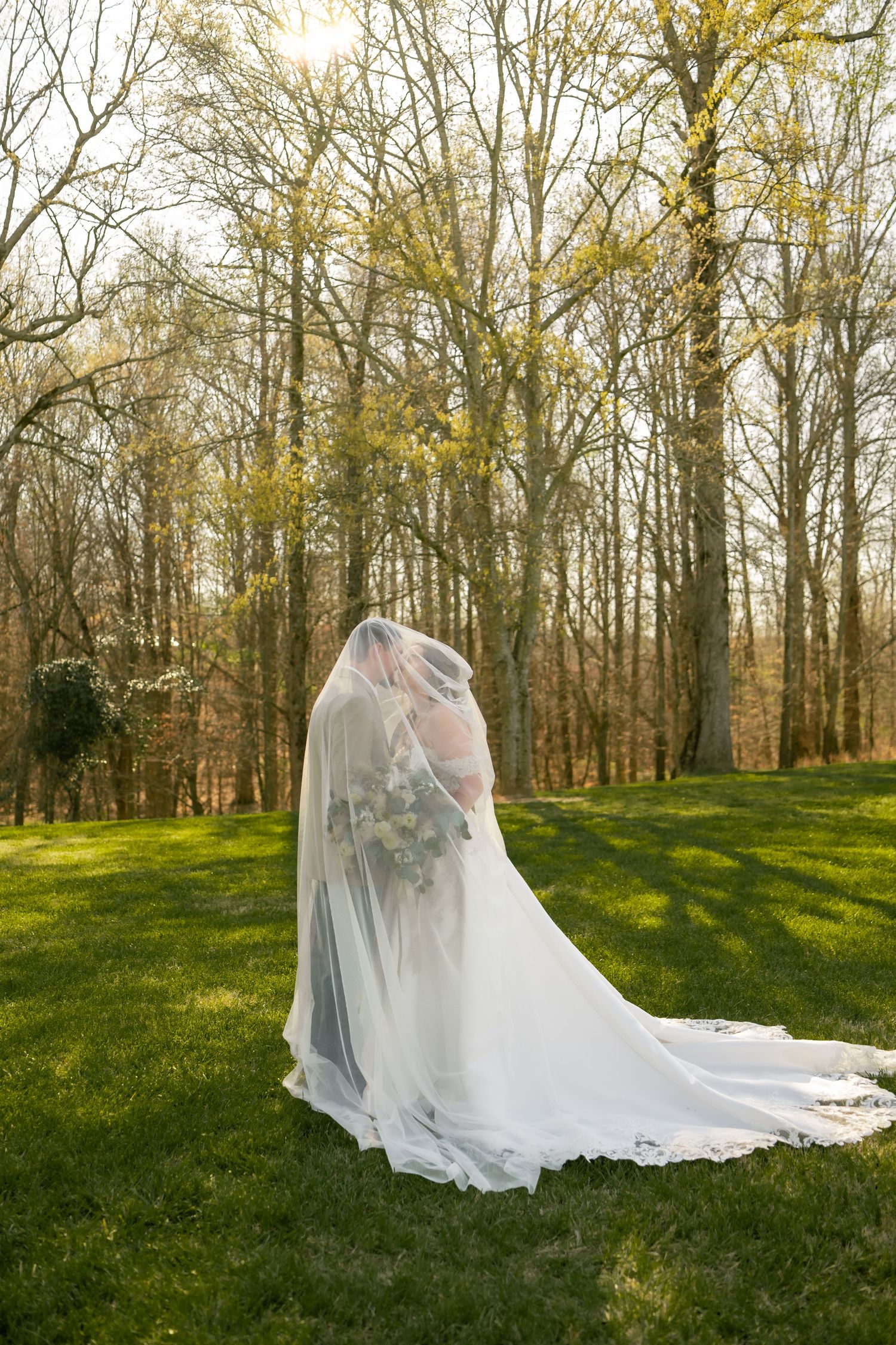 Bride in white gown and long veil stands on green grass surrounded by tall trees in an outdoor woodland setting.