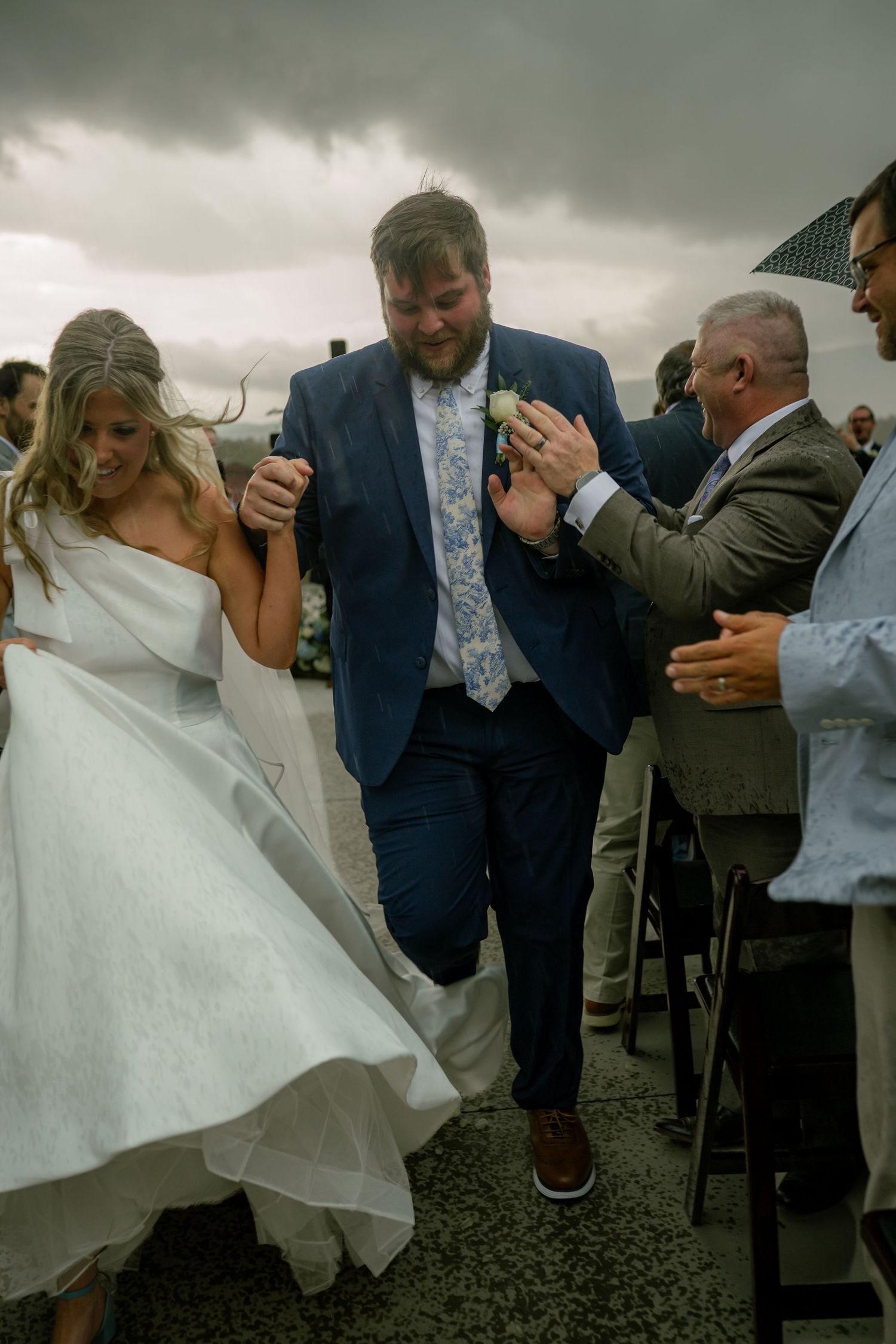 Bride and groom dash through cheering guests under stormy skies during outdoor wedding exit.