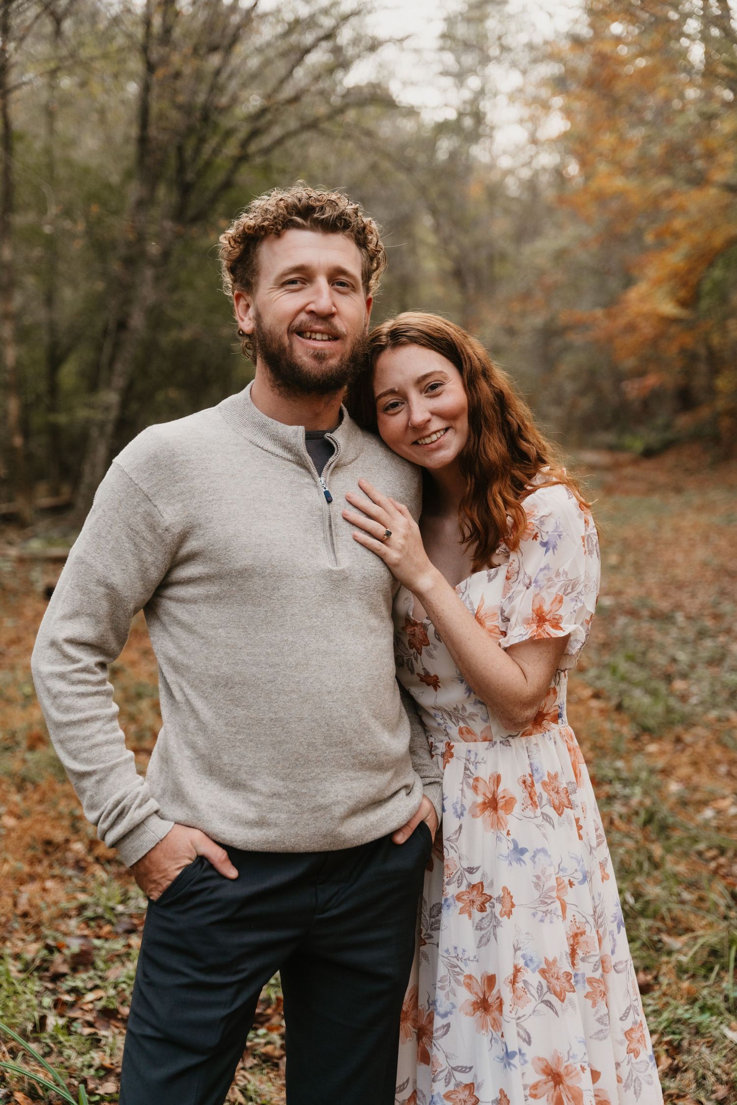 A romantic autumn engagement photo shows a couple posing together in a wooded setting with fallen leaves.