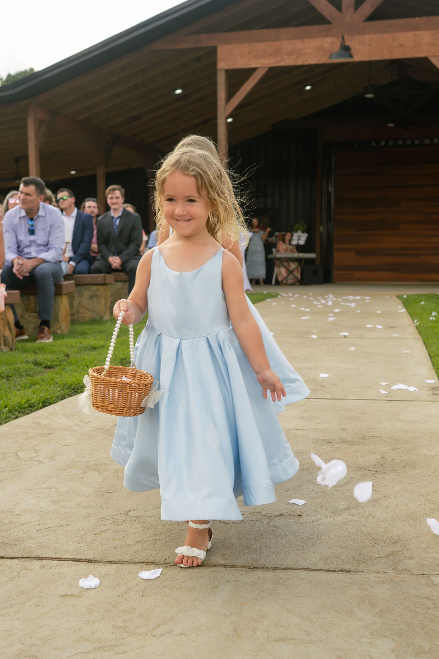 Adorable flower girl in light blue dress scatters white petals down outdoor wedding aisle while smiling joyfully.