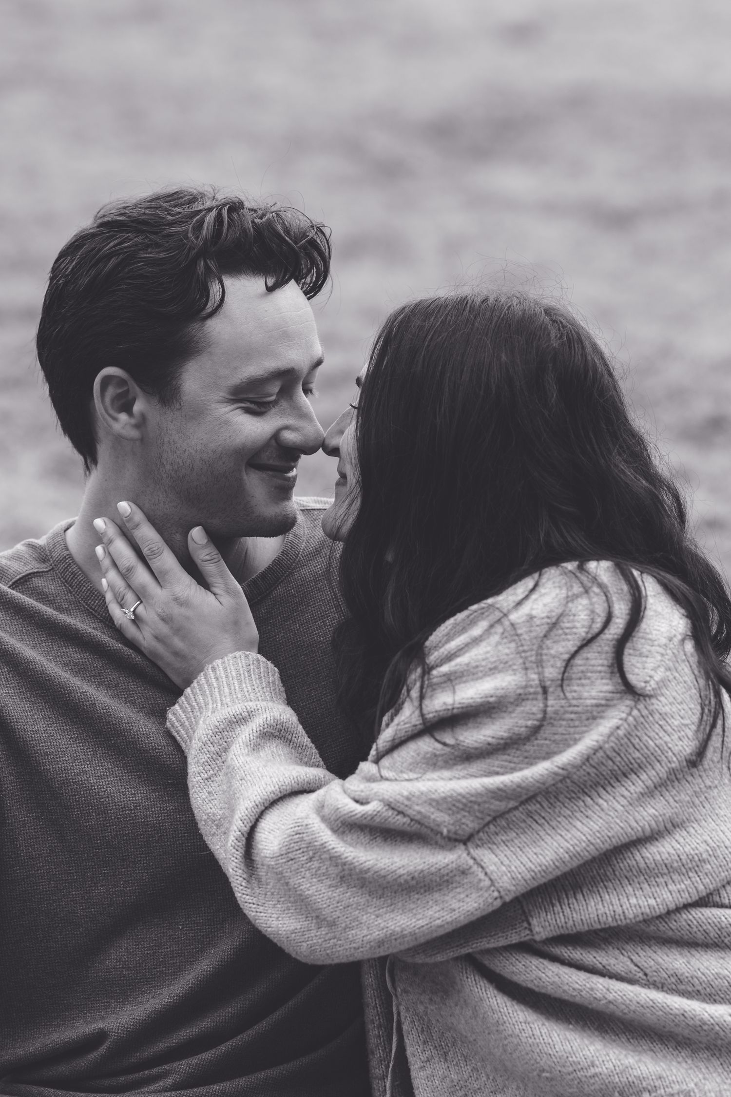 Black and white photo of a smiling couple in sweaters sharing a tender romantic moment outdoors.