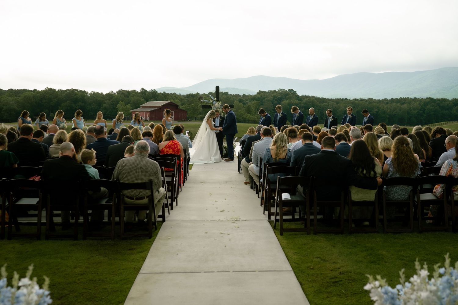 Outdoor wedding ceremony with guests seated along white aisle, couple at altar, mountain backdrop under cloudy sky.