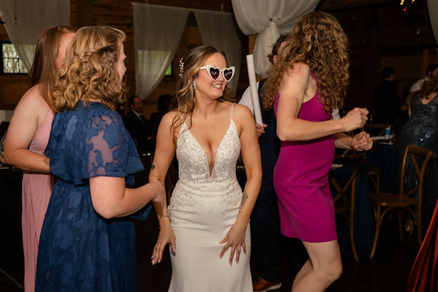 Bride in white gown wearing heart-shaped sunglasses dancing joyfully with guests at lively wedding reception.