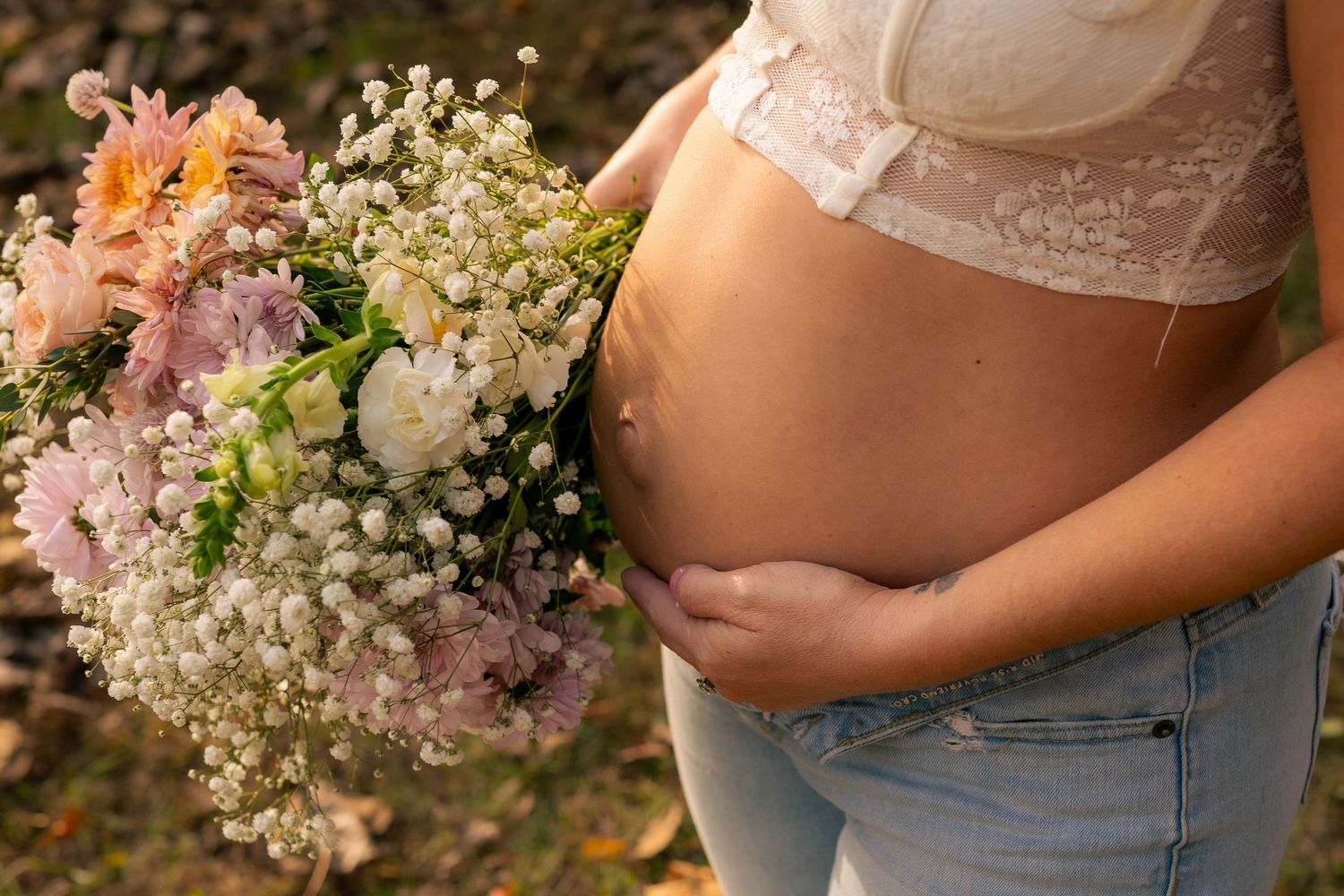 A pregnant belly in lace and denim holds a delicate bouquet of baby's breath, gerbera daisies and pink flowers.