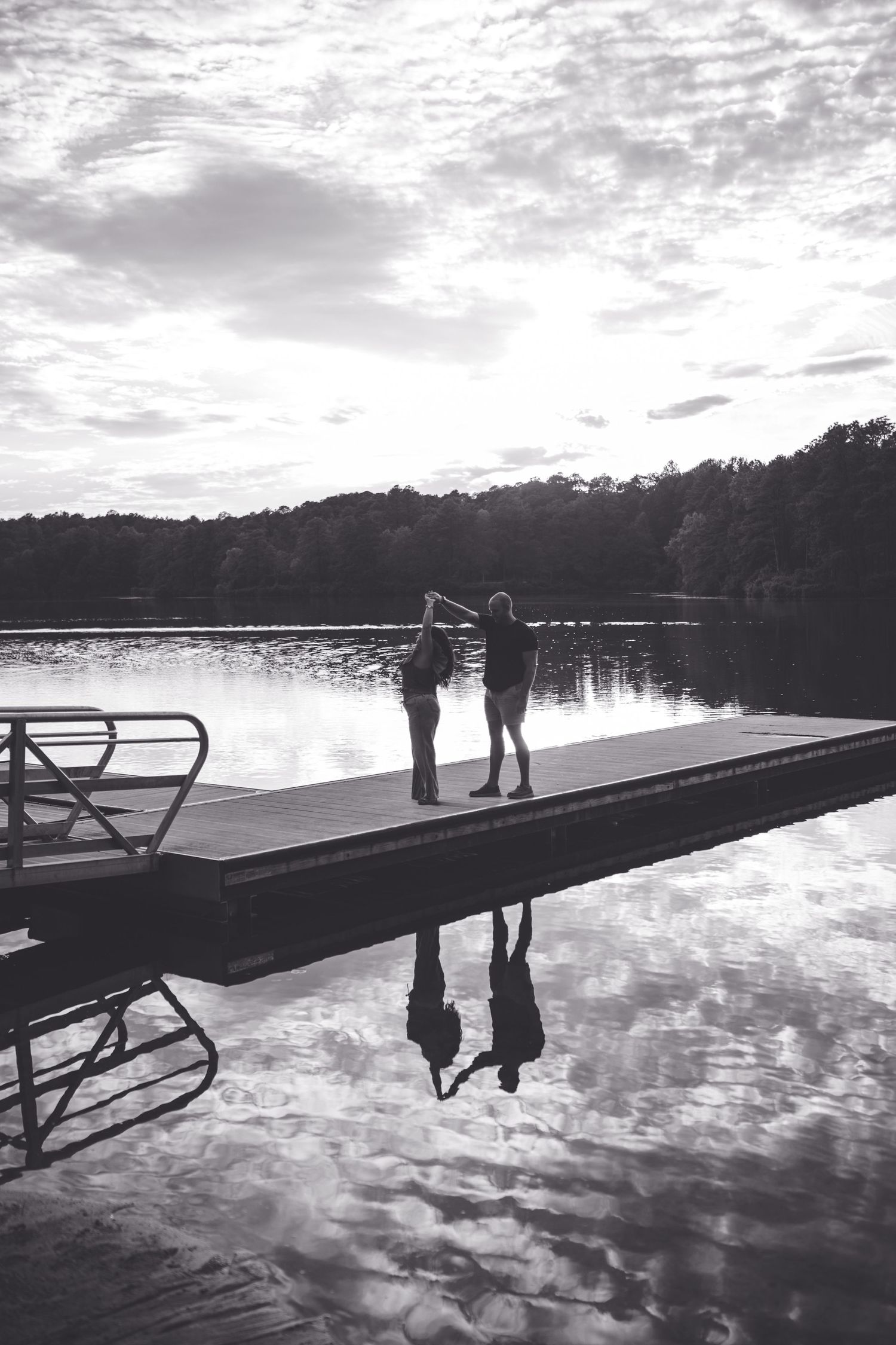 Two people stand on a lake dock in black and white, their reflections mirrored in calm water with trees behind.