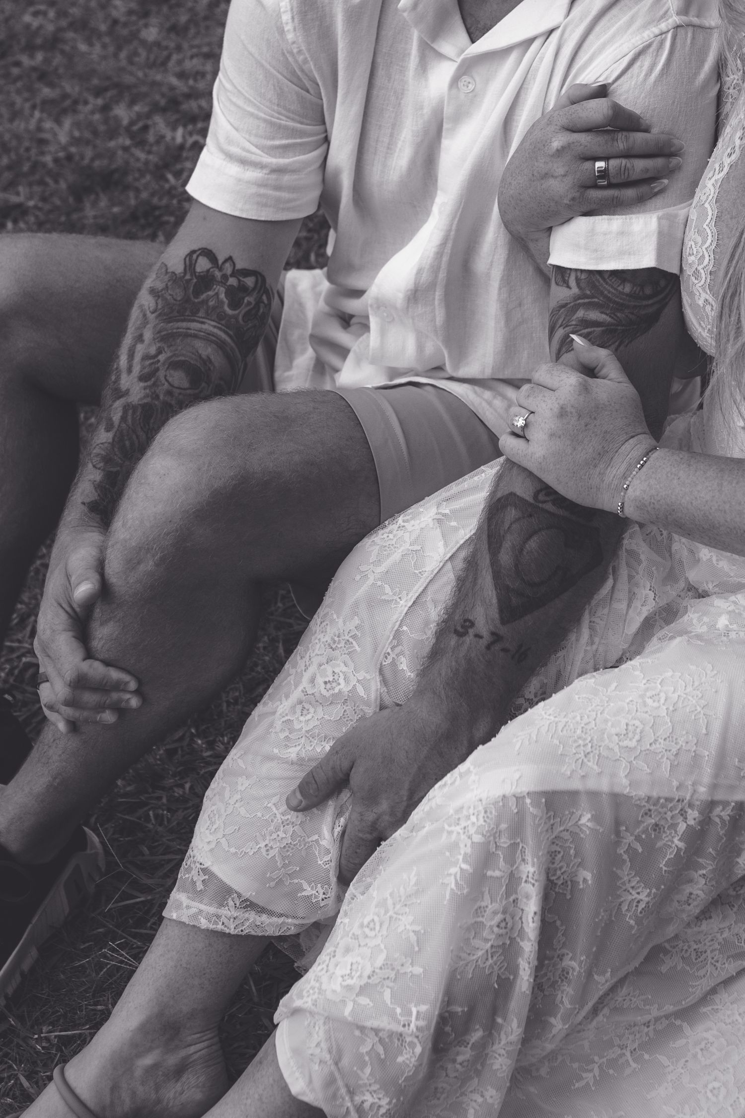 Black and white photo of tattooed couple sitting together, wearing wedding rings, in casual white attire.