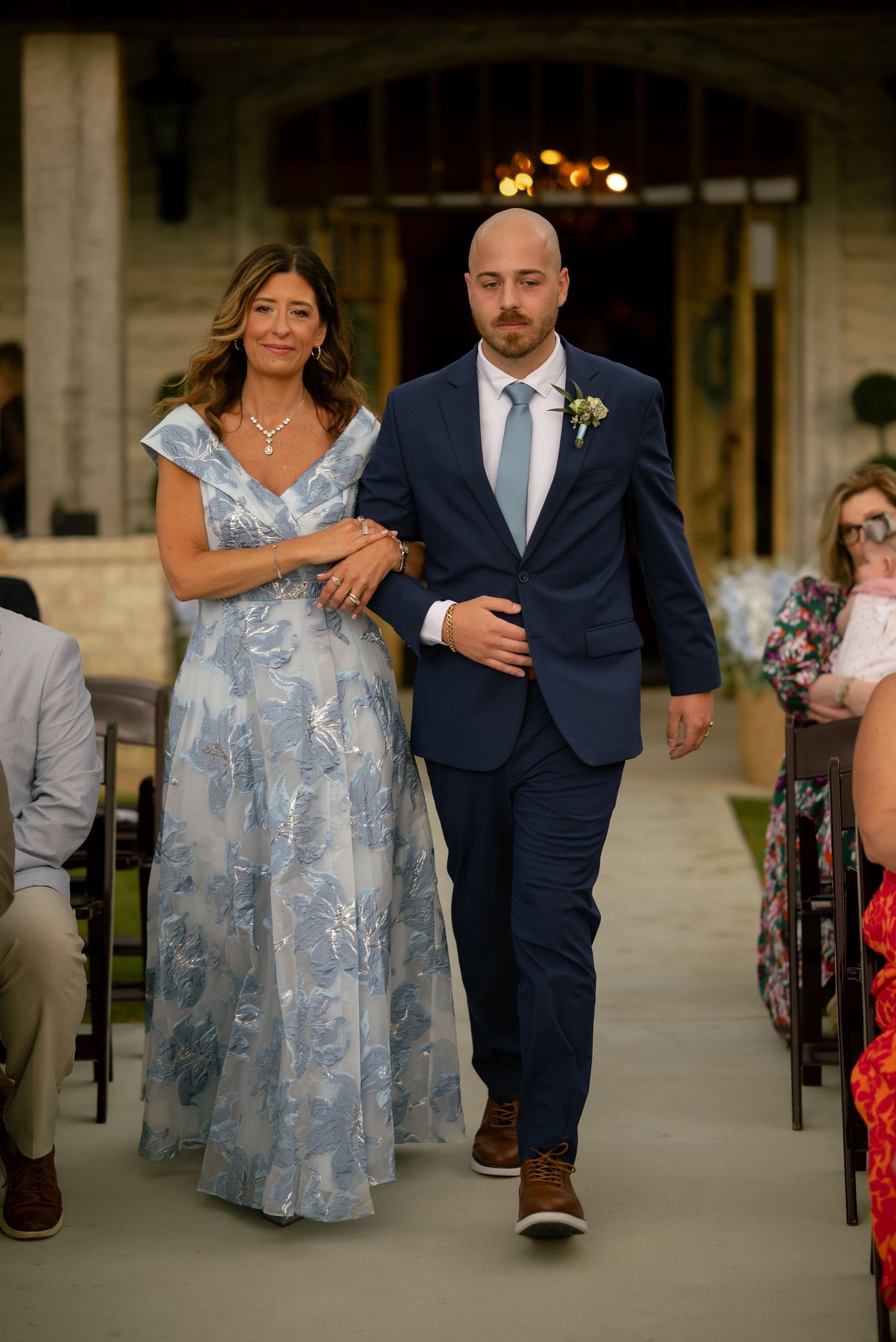 Groom in navy suit escorts woman in blue floral gown down wedding ceremony aisle.