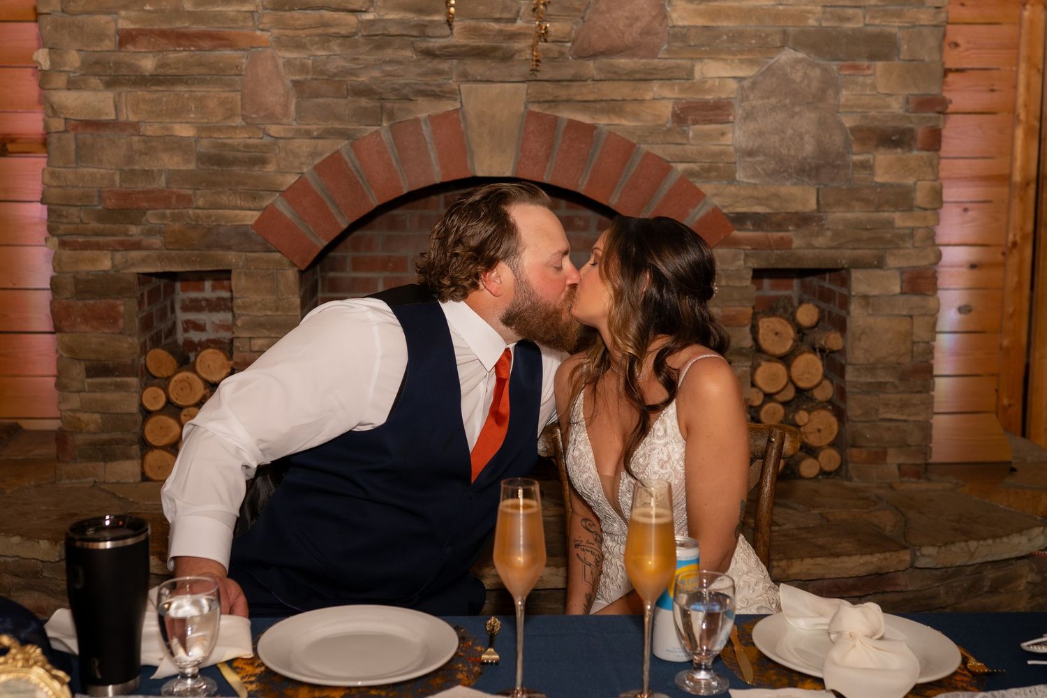 Bride and groom share a kiss at sweetheart table in front of stone fireplace during wedding reception.