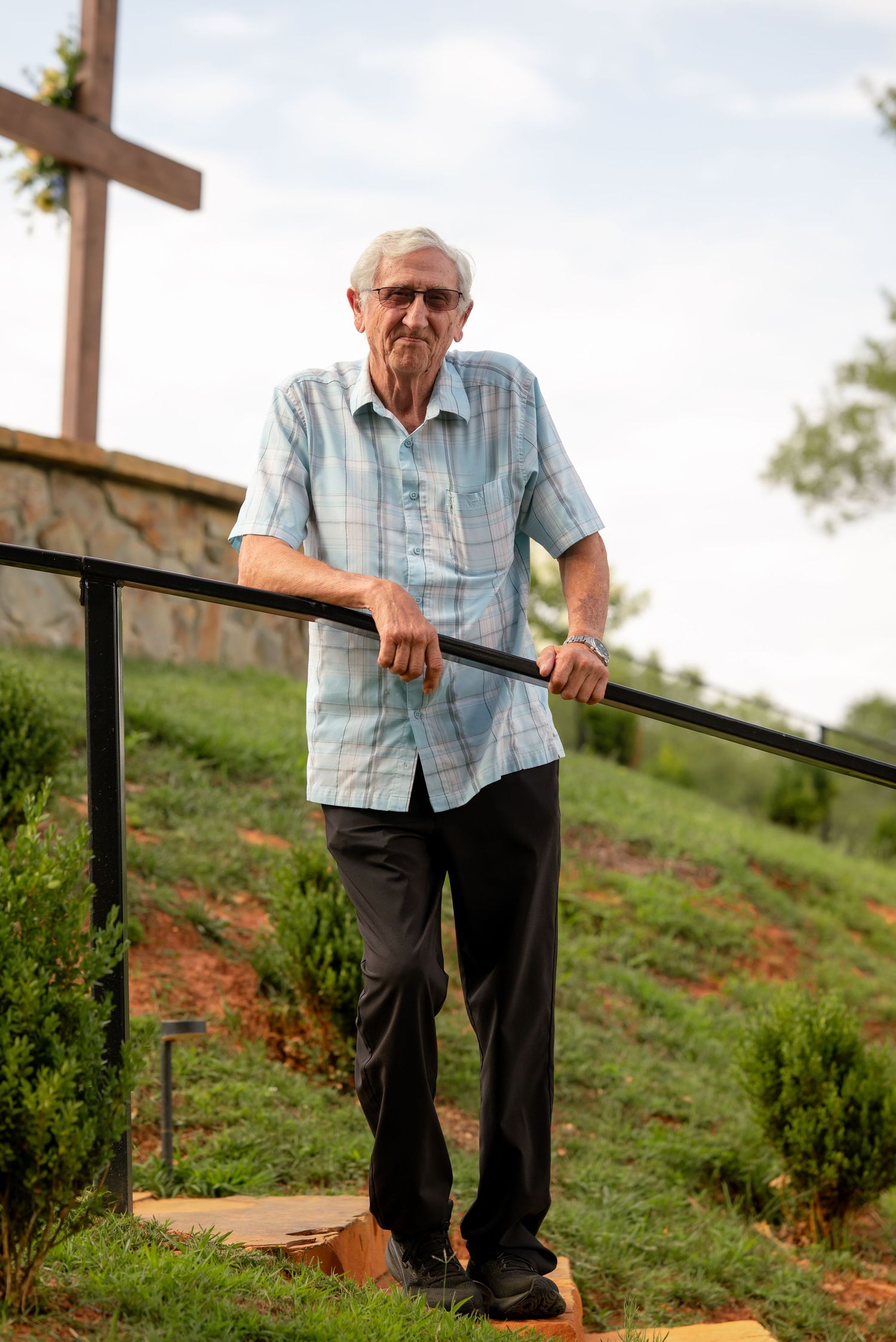 Elderly man in plaid shirt stands on outdoor steps holding black railing near large wooden cross on grassy hill.