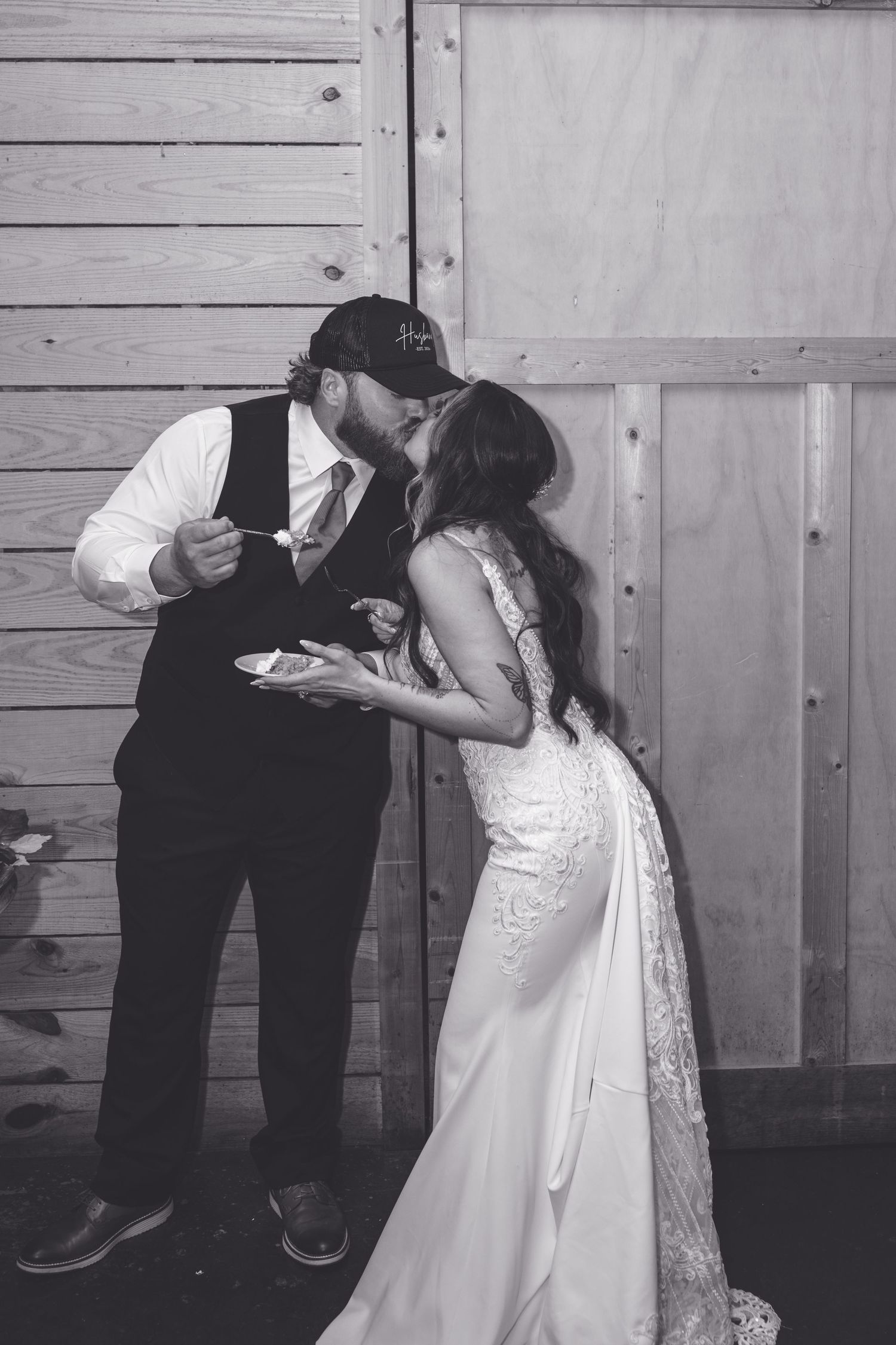 Bride and groom share a kiss while cutting wedding cake against wooden barn wall in black and white photo.