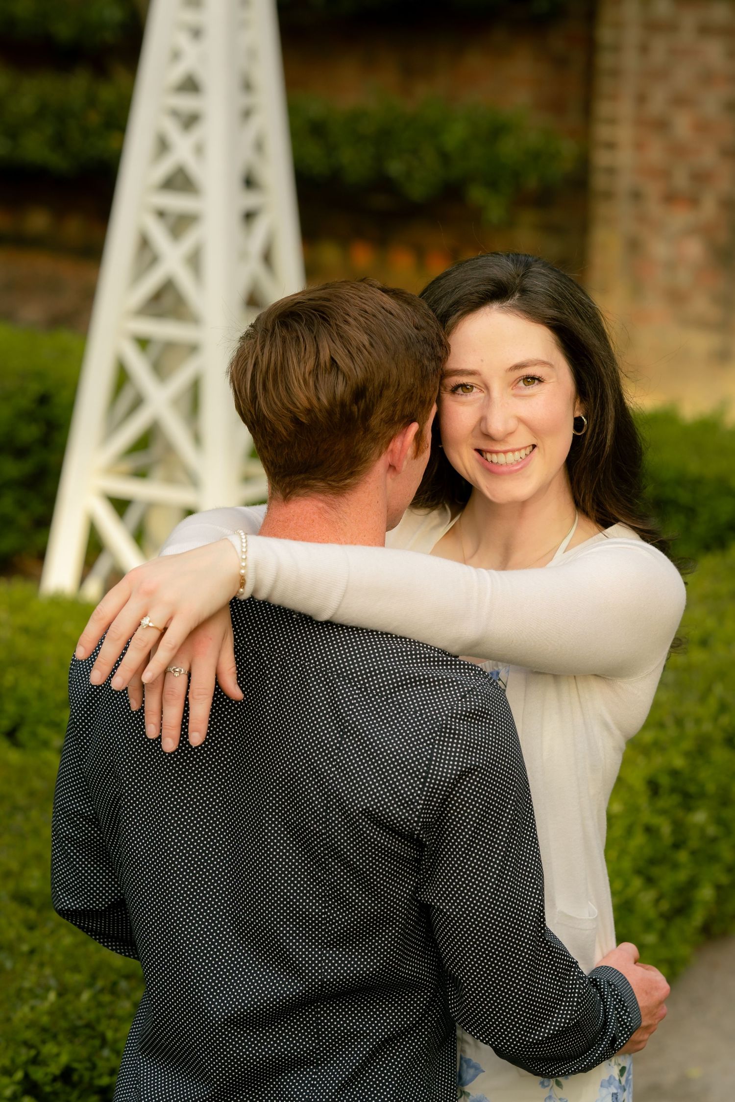 A smiling brunette woman embraces a man outdoors near green hedges and a white decorative windmill.