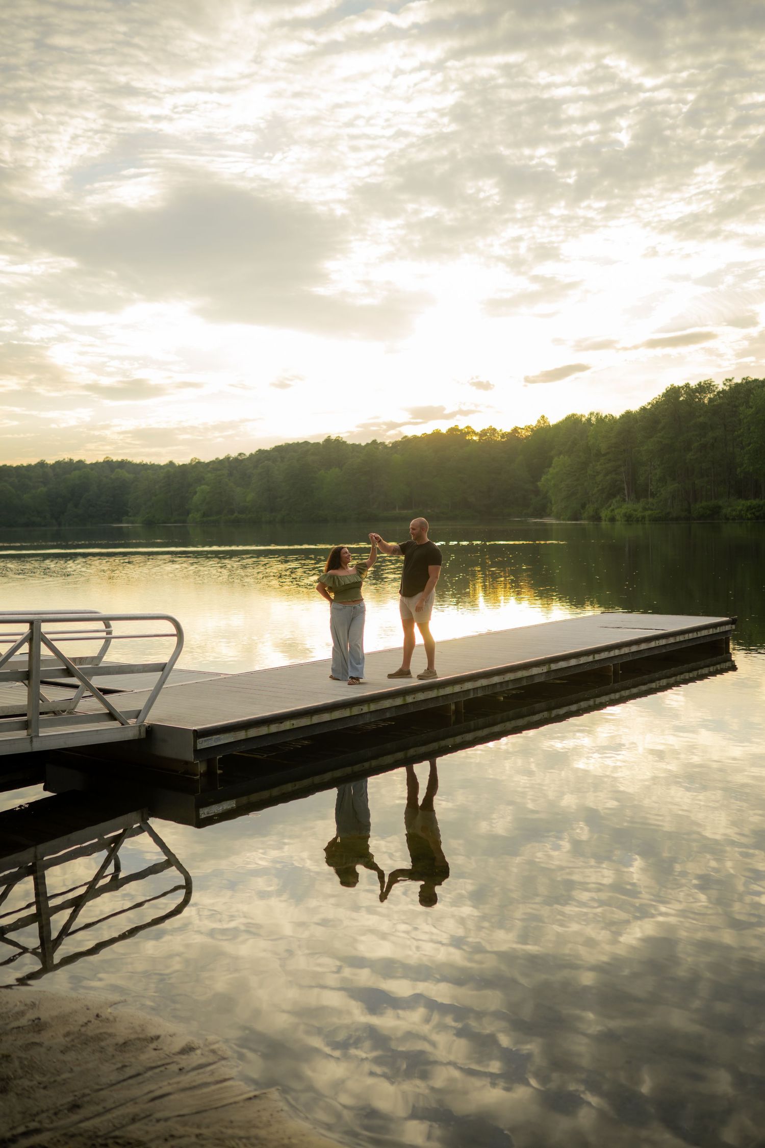 Two people stand on a calm lake dock at golden sunset, their reflections mirrored in still water with forested hills behind.