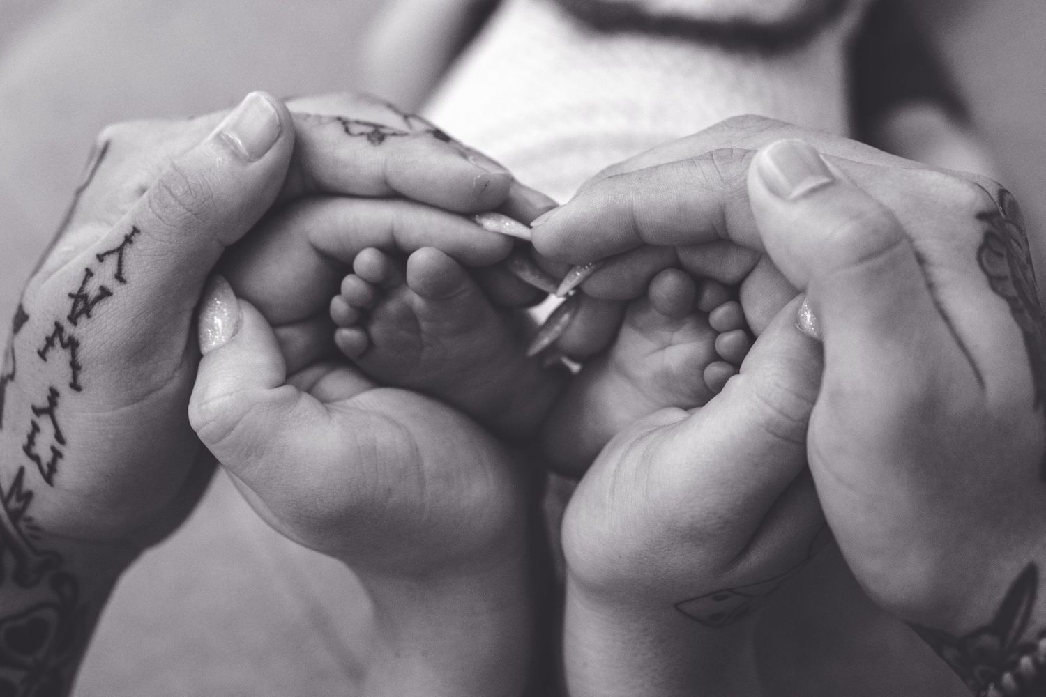 Black and white image of tiny baby feet cradled in tattooed adult hands forming a heart shape.