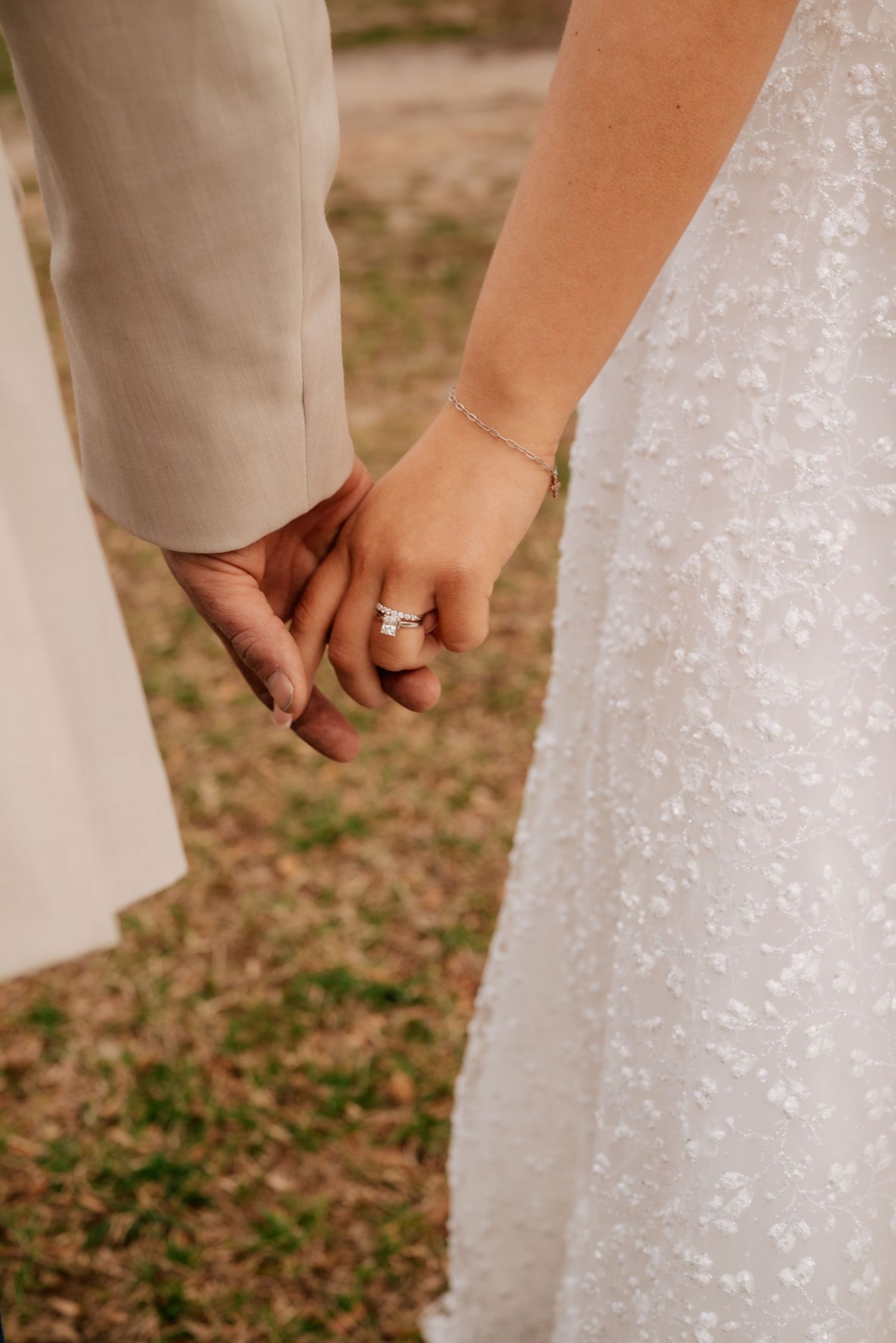 Close-up of newlywed couple holding hands, showing engagement ring and white sequined wedding dress against outdoor setting.