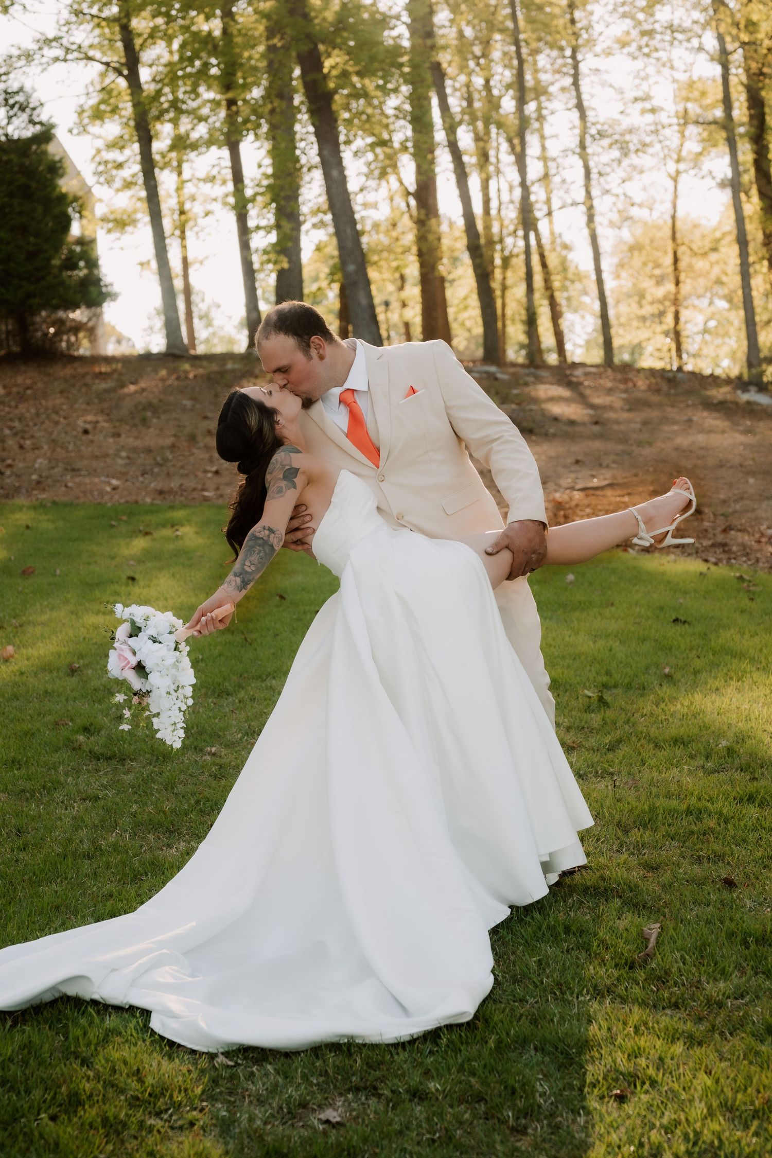 A romantic dip pose of newlyweds on a grassy lawn with sunlit trees in the background as the bride holds a white bouquet.
