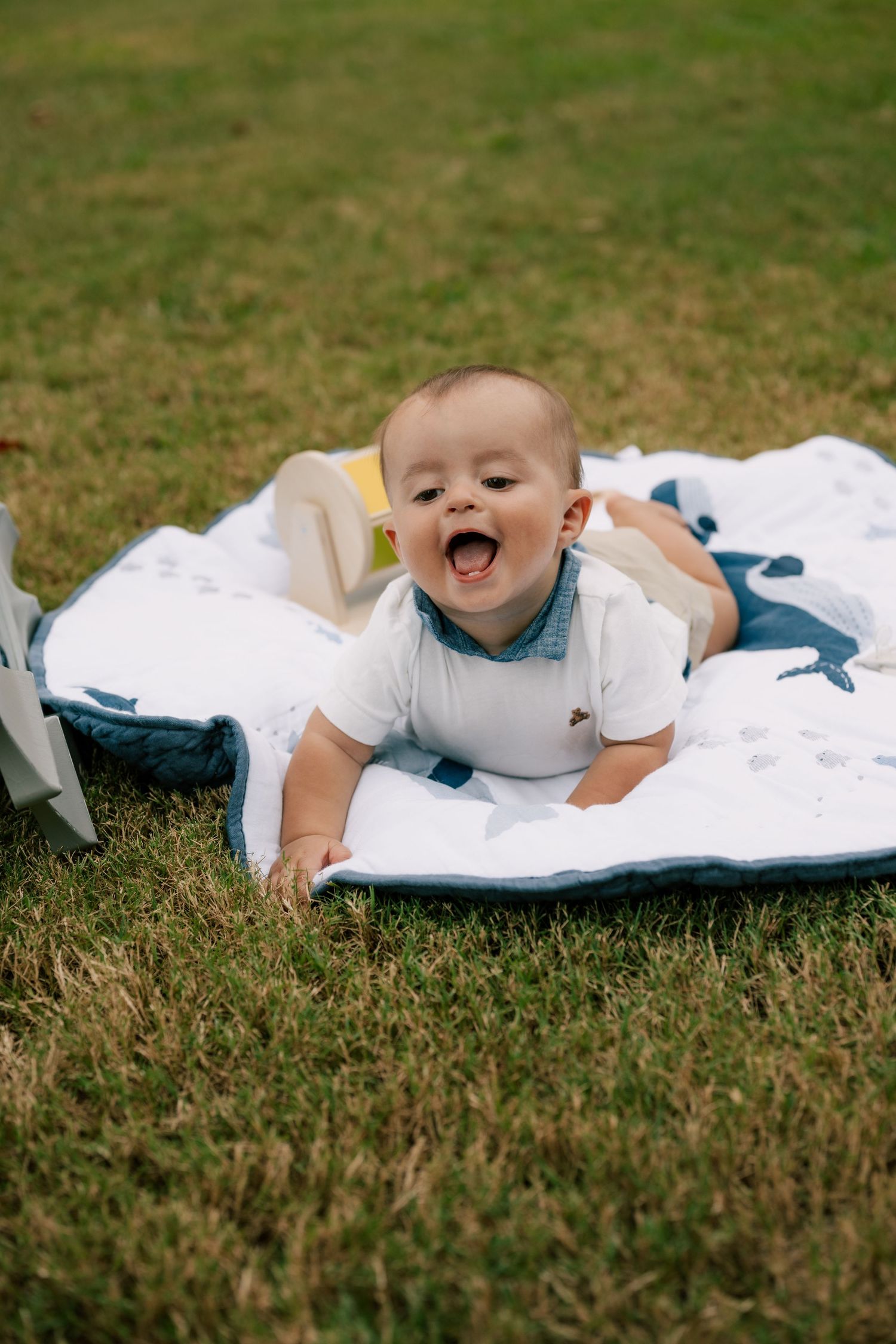 A baby lies on a white blanket on grass while laughing and playing outdoors.