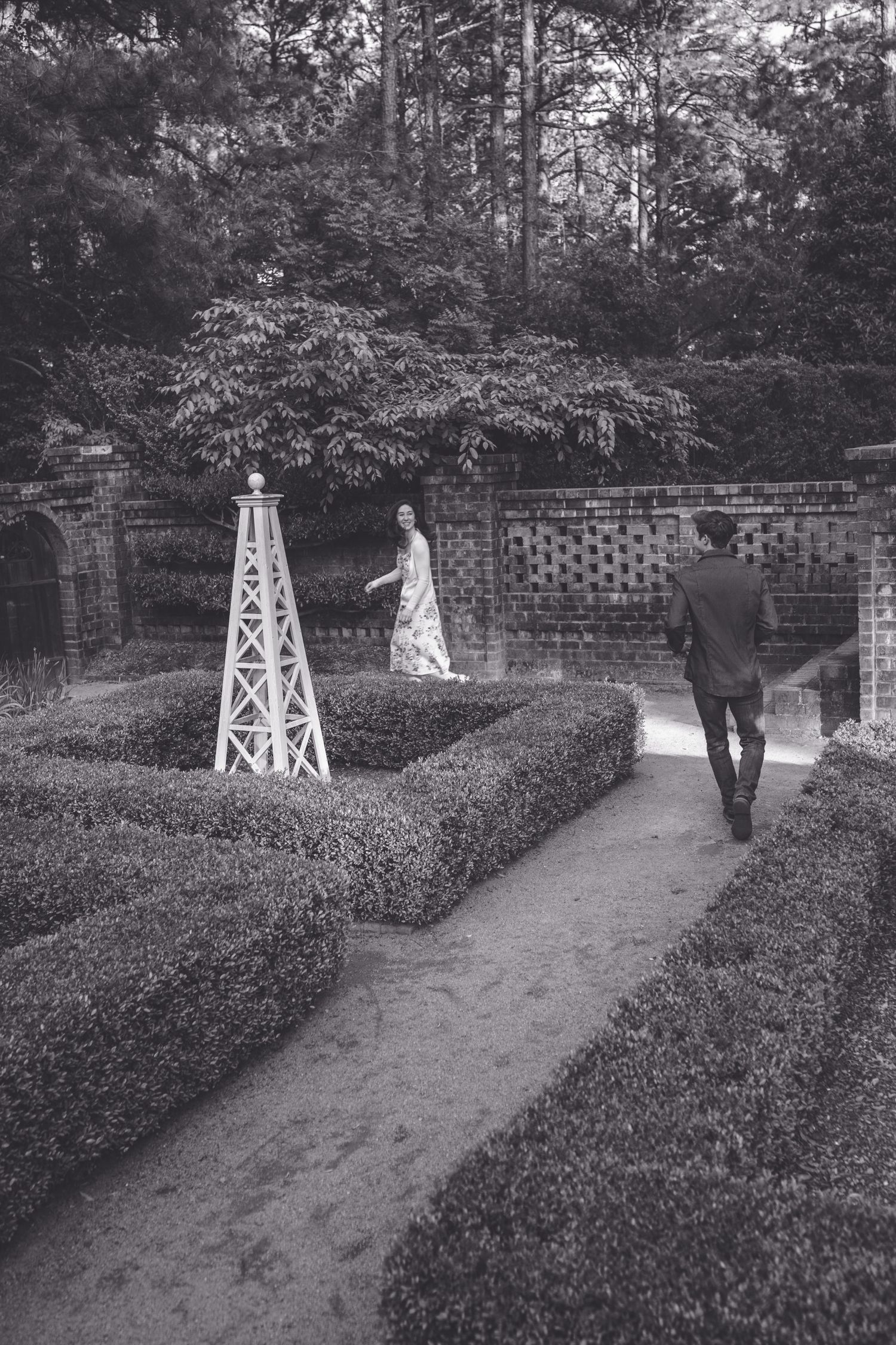 Black and white photo of a bride and groom walking through a formal garden maze with brick walls and hedgerows.