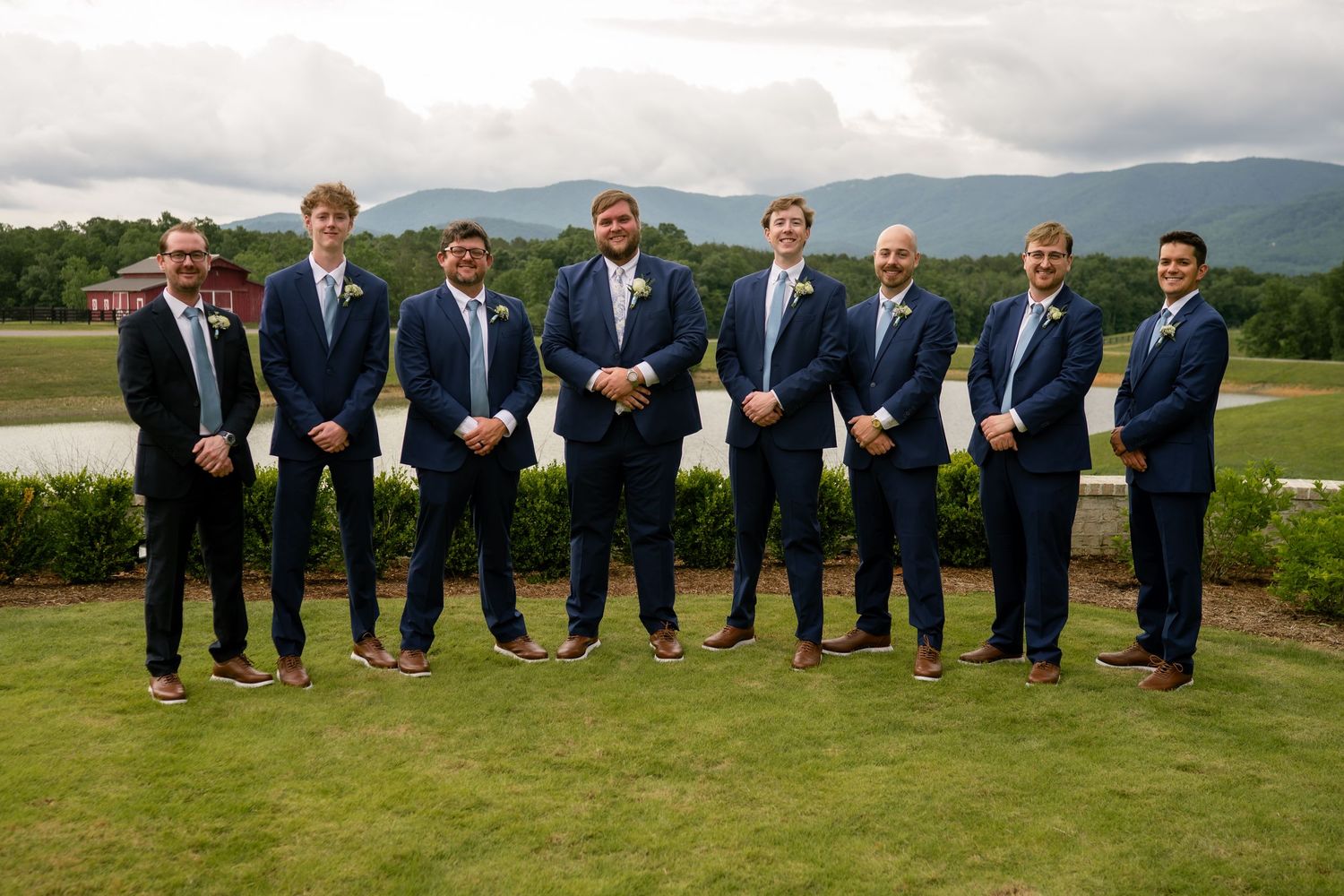 Eight groomsmen in navy suits pose outdoors at a wedding venue with mountain views and lush greenery.