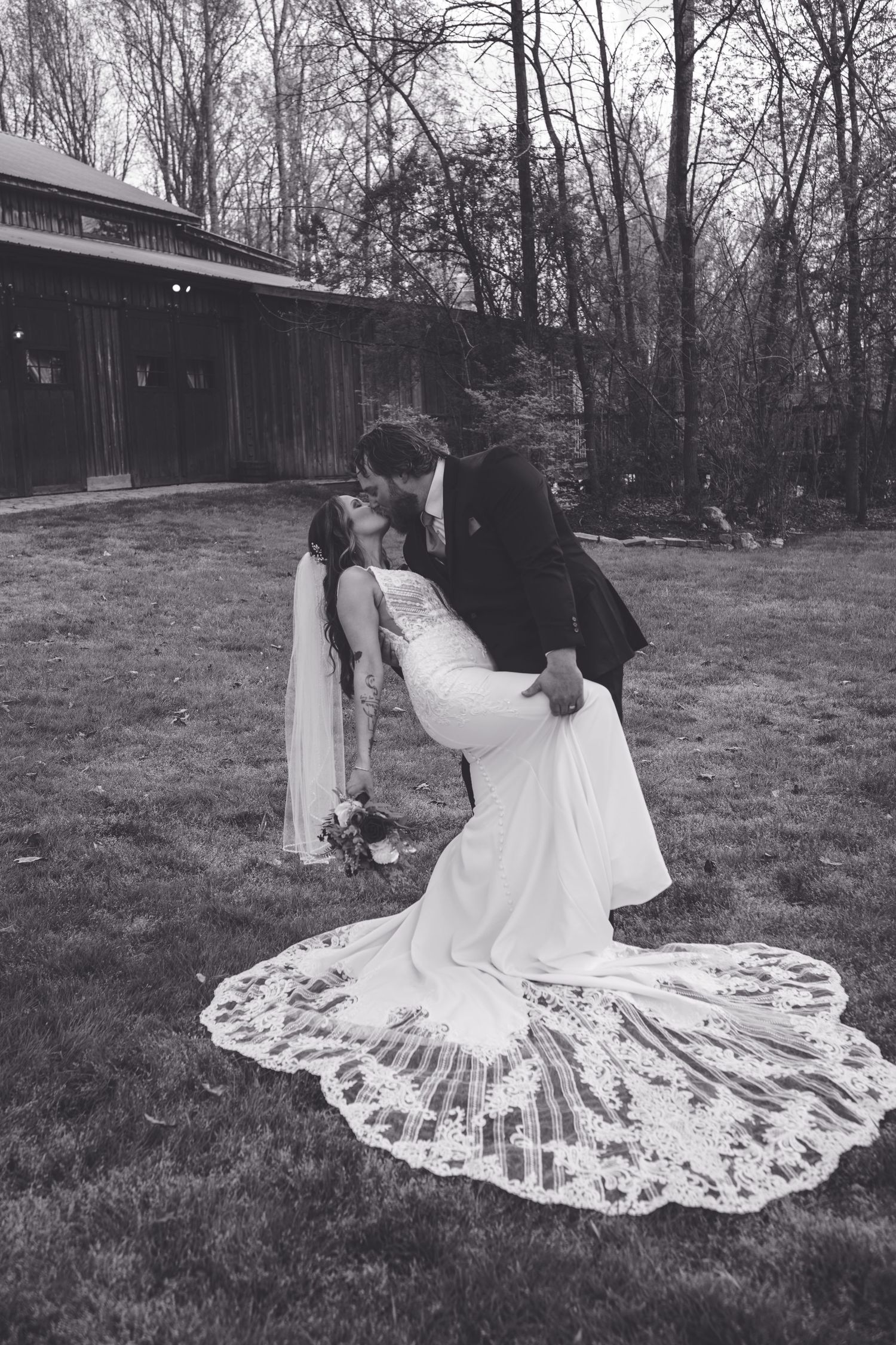 Black and white photo of groom dipping bride for romantic kiss outdoors after wedding ceremony.