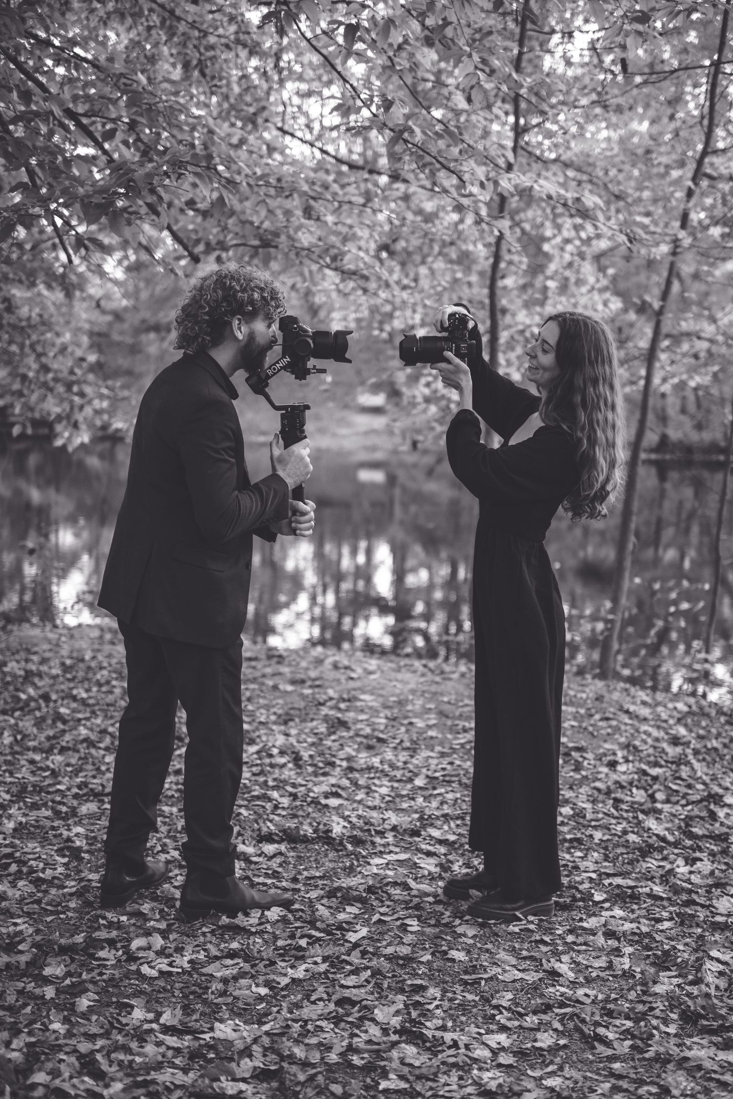 Two photographers stand in a wooded area by a lake, pointing cameras at each other in black and white. Southern Pines, NC