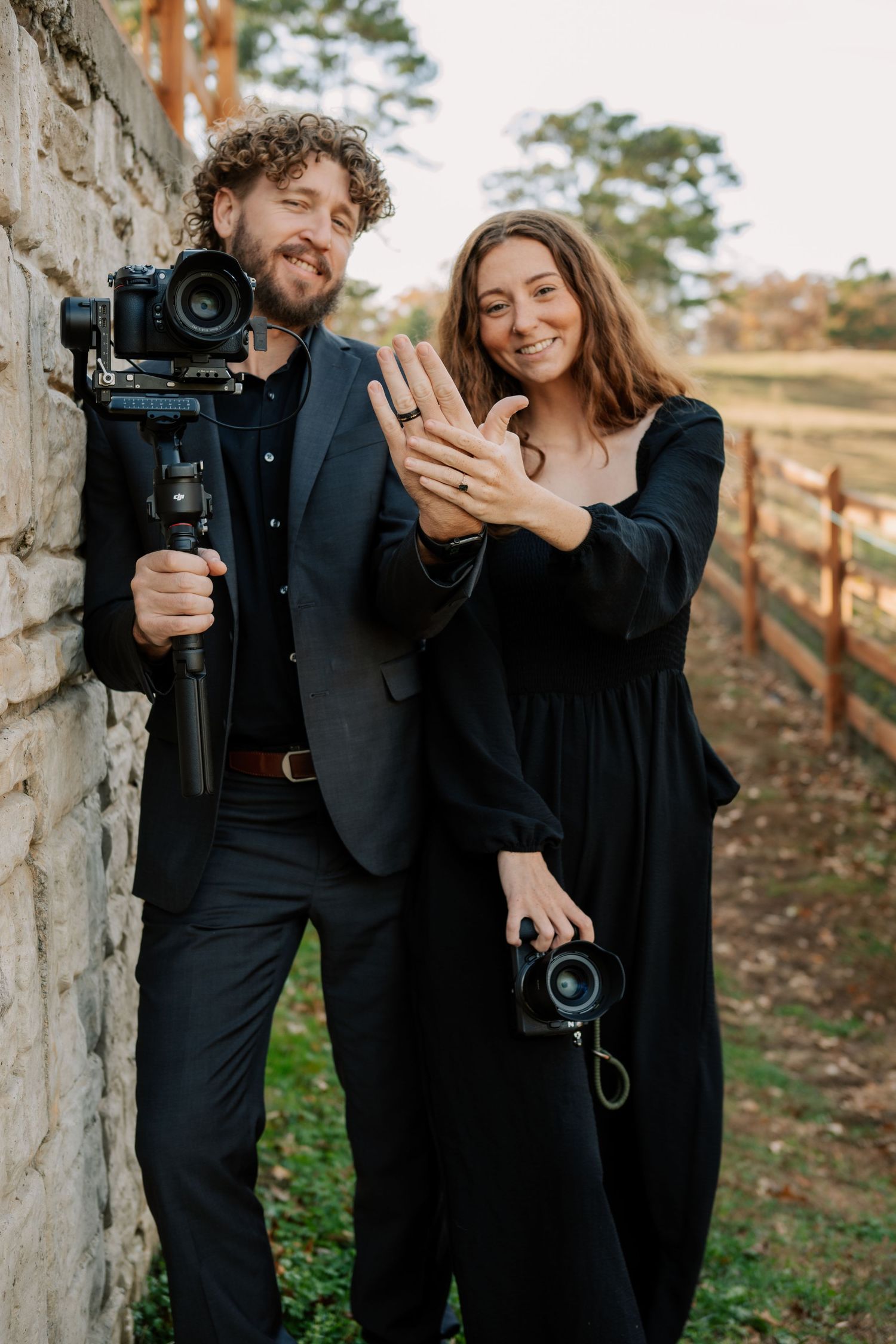 A couple in black outfits laughs together while holding professional cameras against a rustic stone wall outdoors.