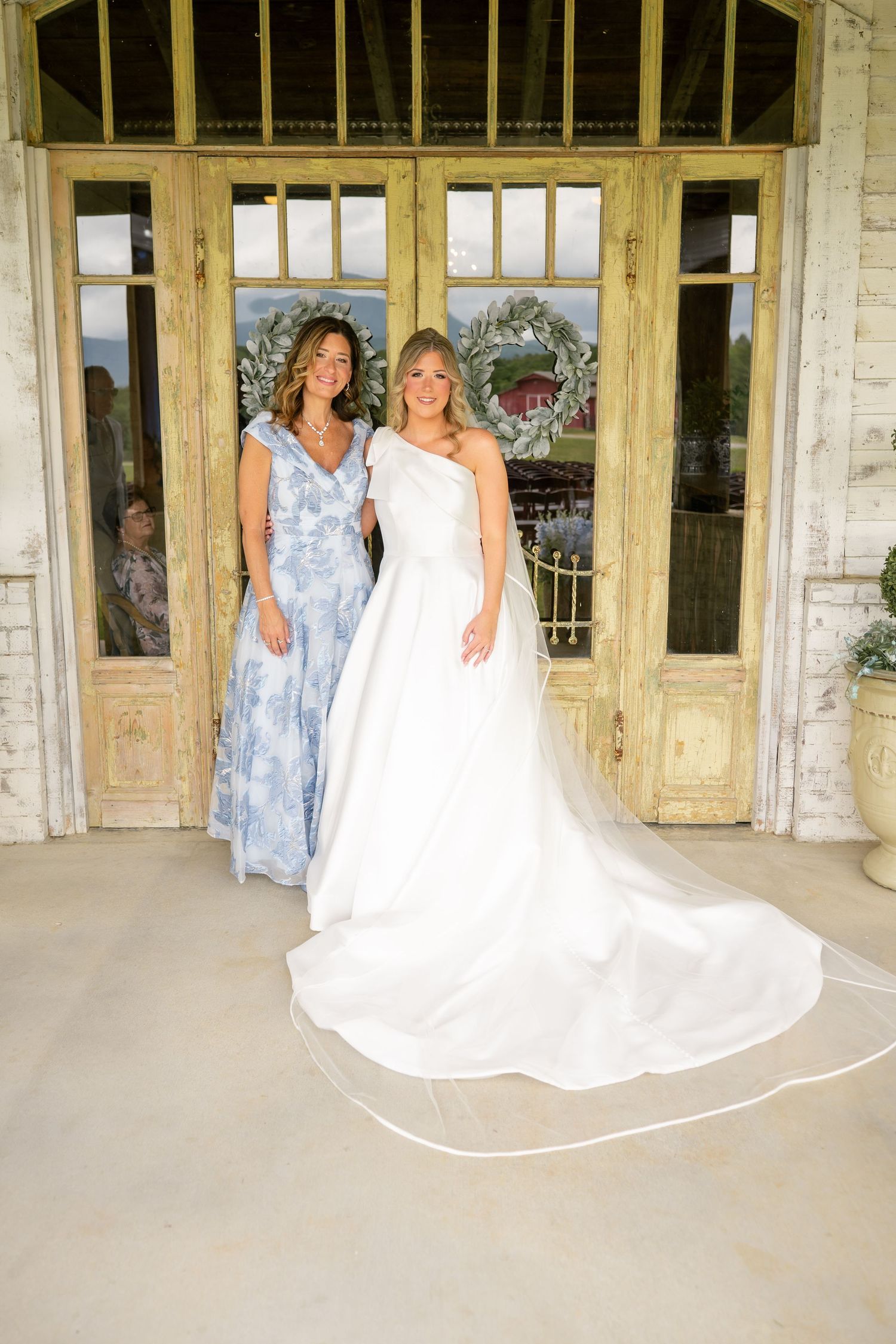 Bride in white one-shoulder gown poses with woman in blue floral dress at venue doors.