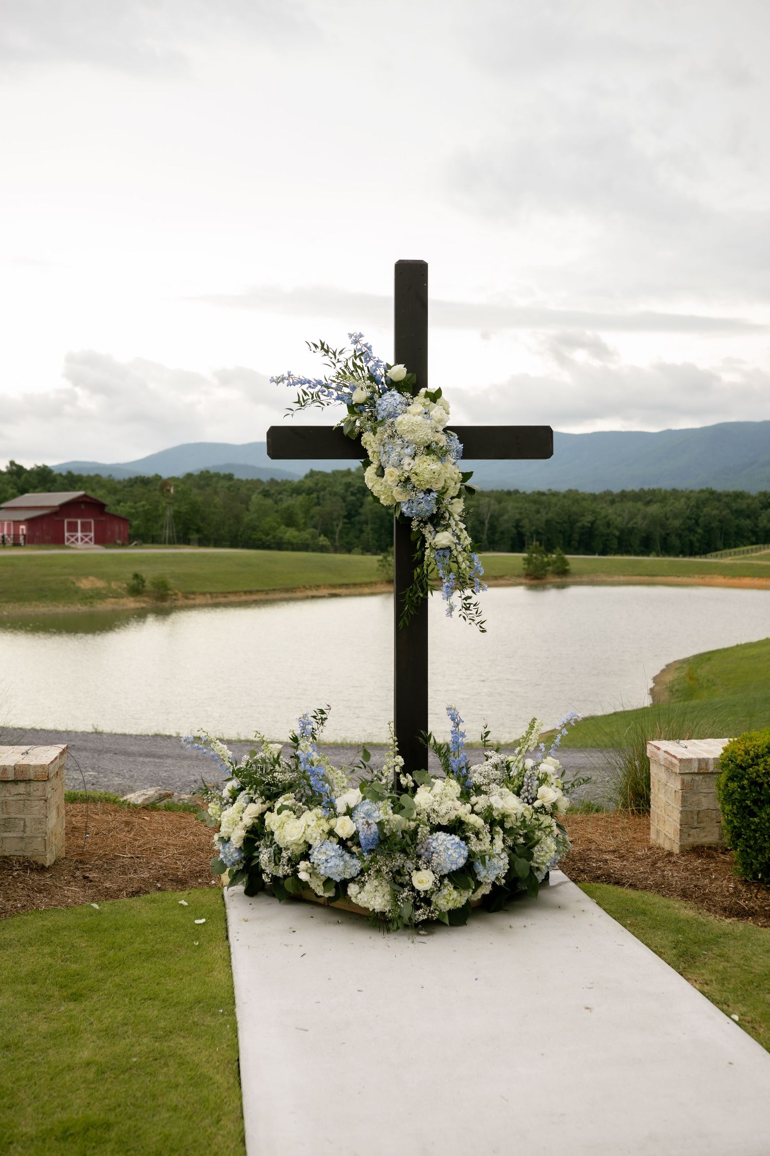 A wooden cross adorned with blue and white floral arrangements stands at an outdoor wedding ceremony beside a serene pond.