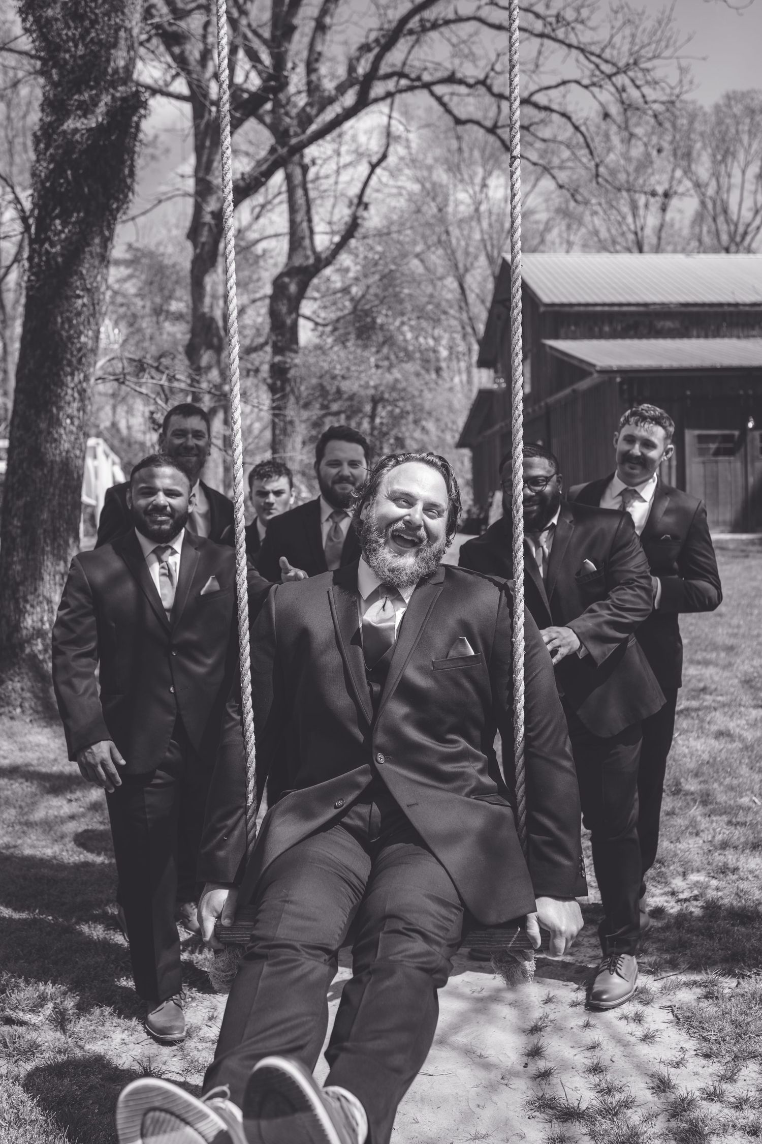 Groomsmen laughing outdoors as groom sits on rope swing in dark suit at rustic wedding venue.